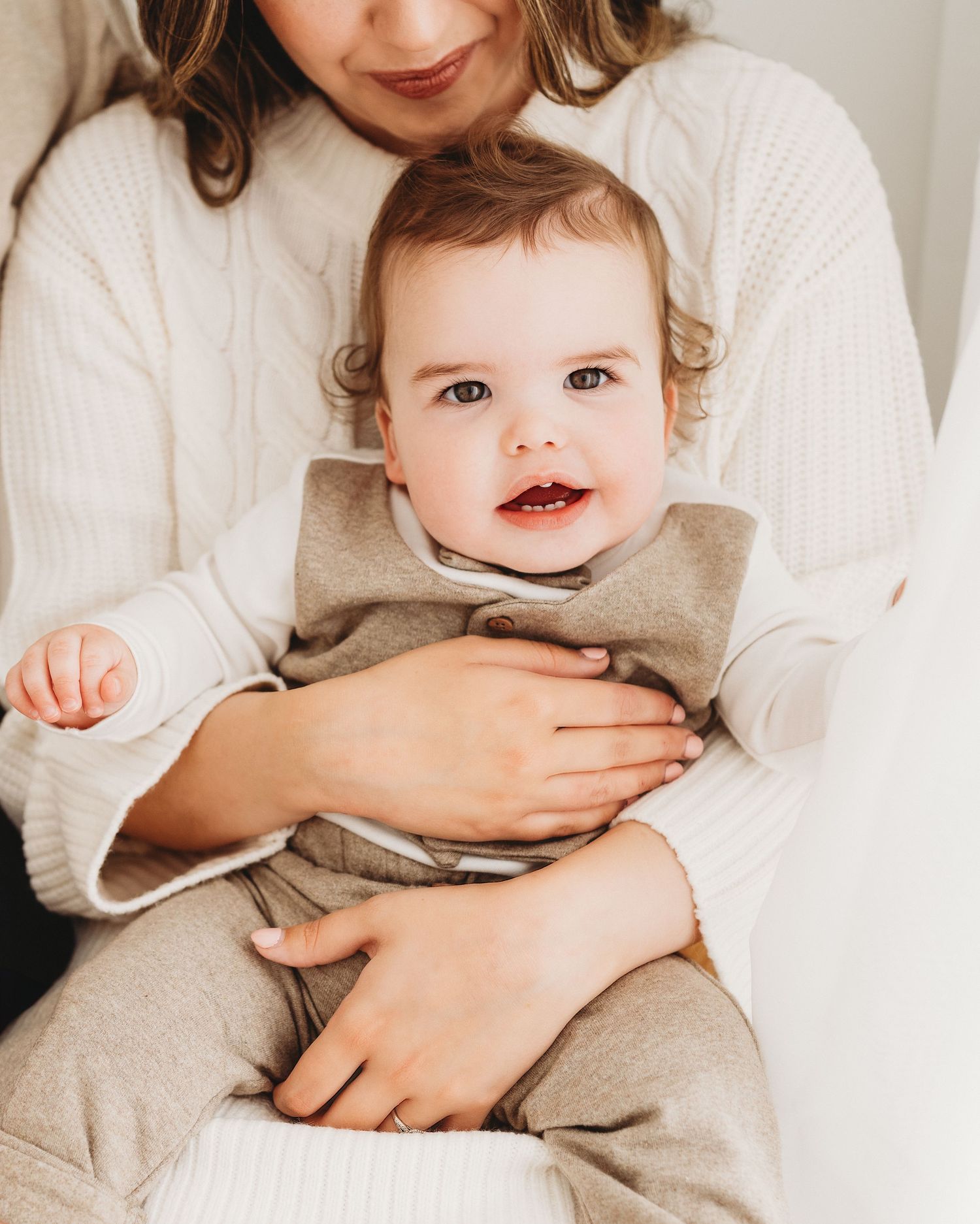 smiling into the camera, 1 year old baby boy sits on mums knee wearing neutral colours during milestone photo session in South Ayrshire