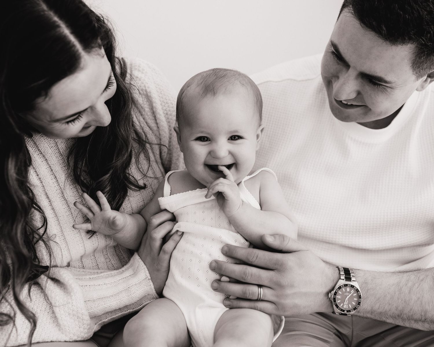 smiling parents look down at giggling baby sat between them during family photoshoot session in Ayrshire studio