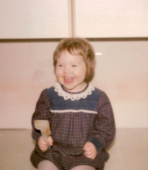 Happy toddler sits smiling captured with cake mix round her face holding the spatula she got to lick