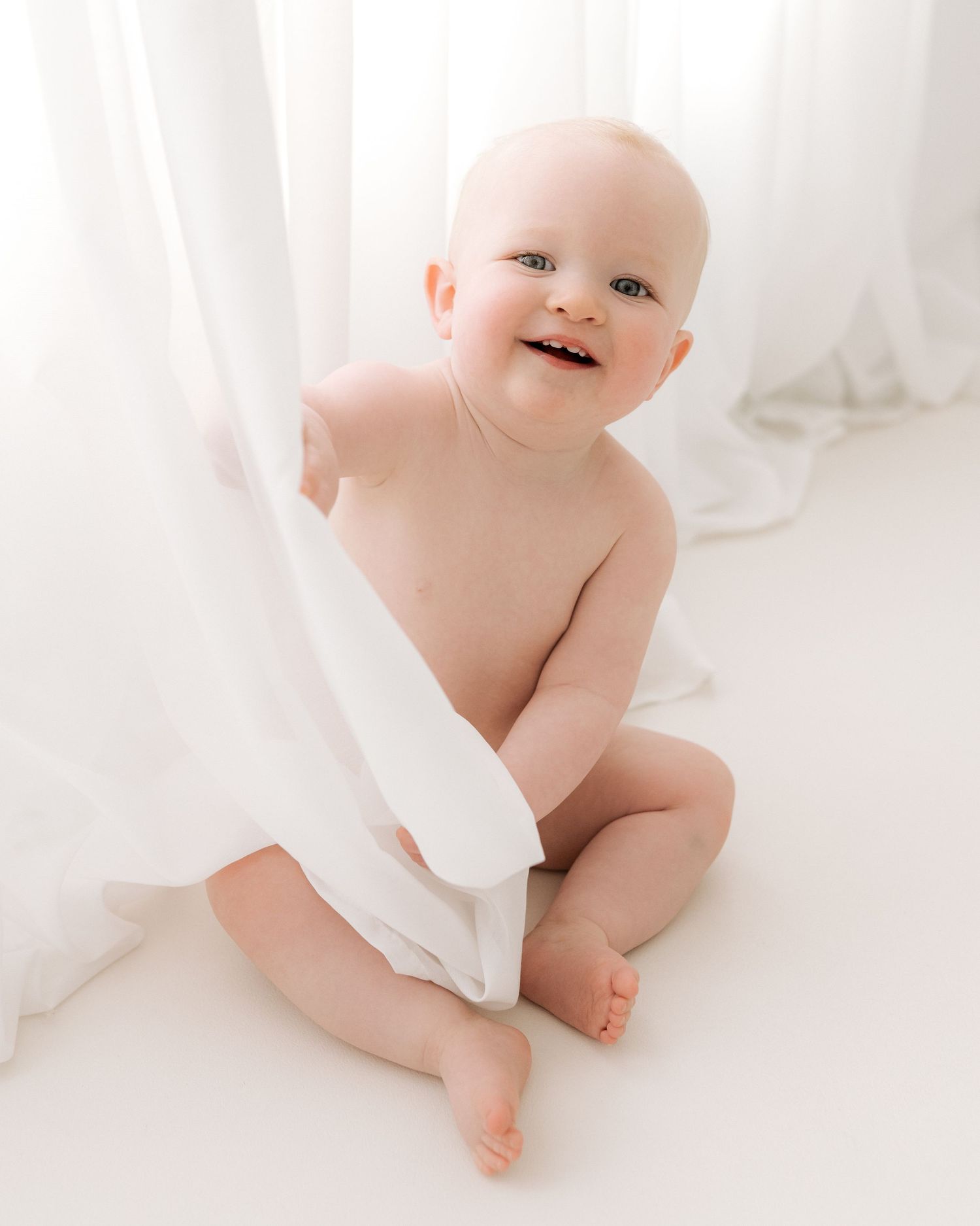 this adorable baby peeks round the curtain she is playing with during her milestone photography session in Ayrshire