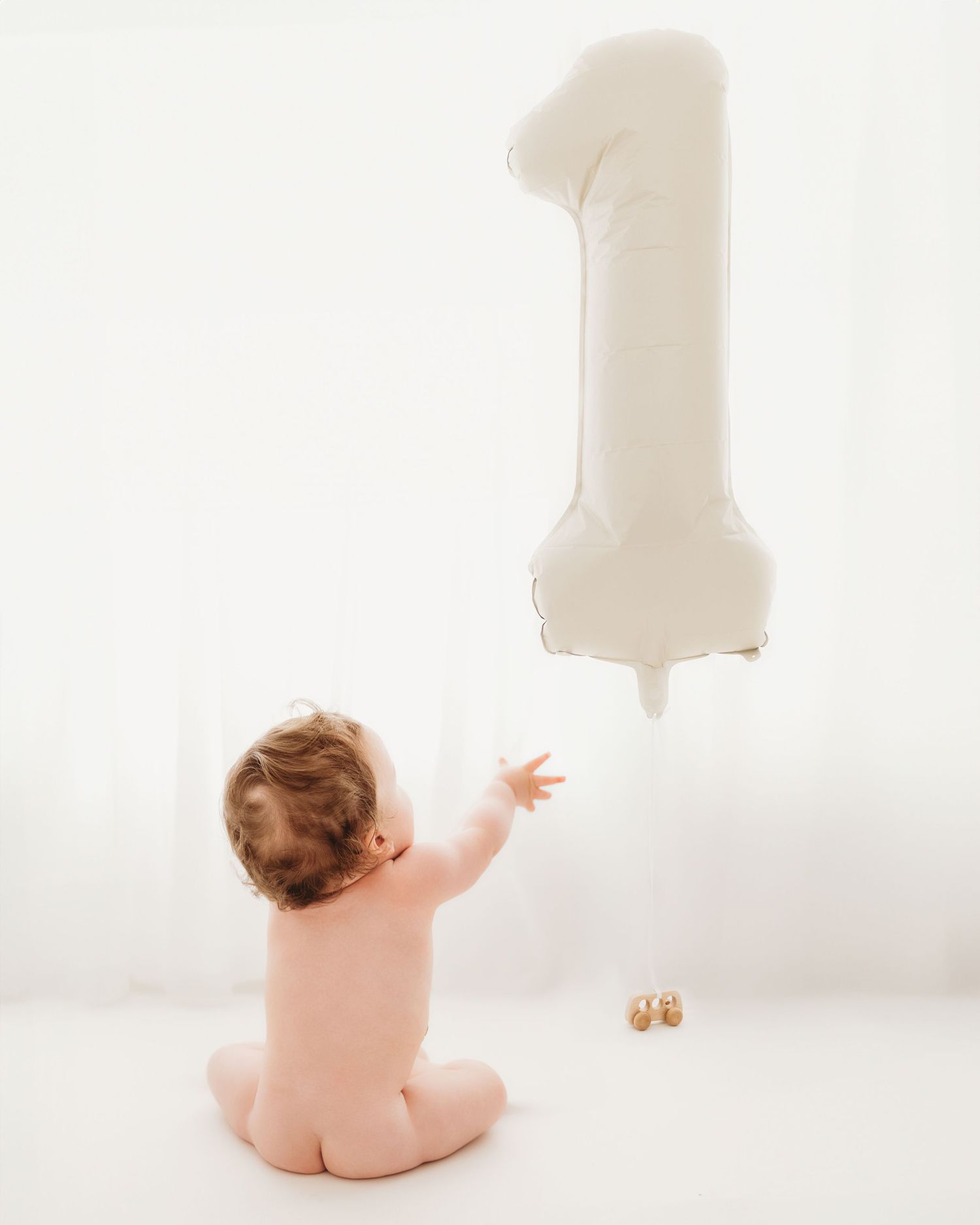 reaching for a number 1 balloon, wearing no clothes, baby celebrates turning 1 during milestone session with his family in Ayrshire studio