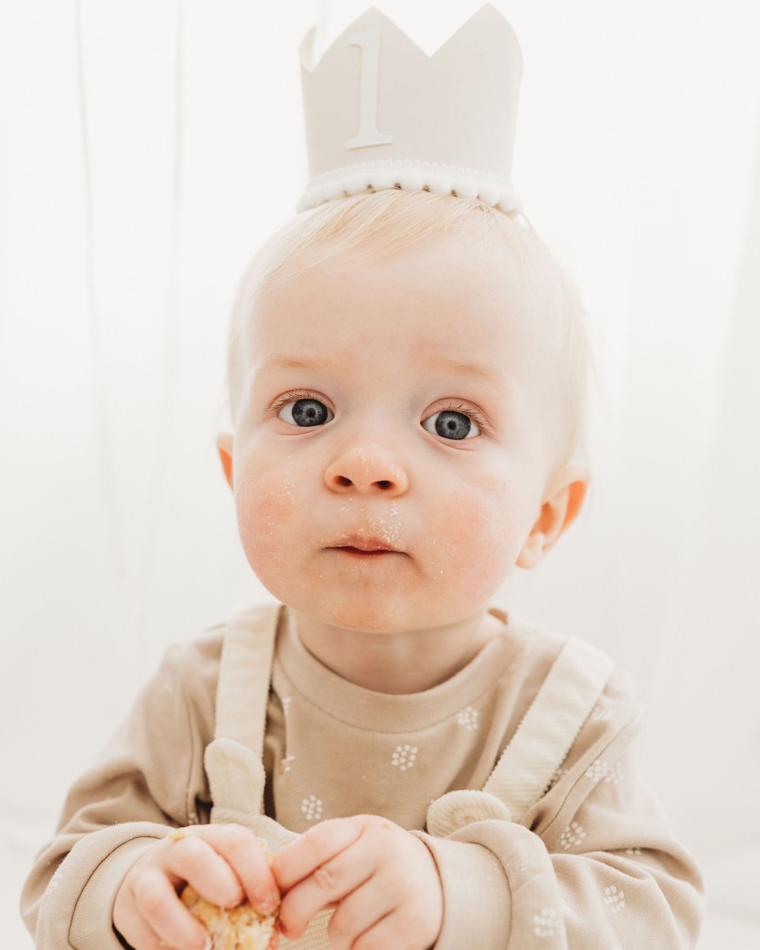 wearing neutral colours and a birthday crown, baby boy looks wide eyed into the camera in Faye McRae's Ayrshire studio during family session