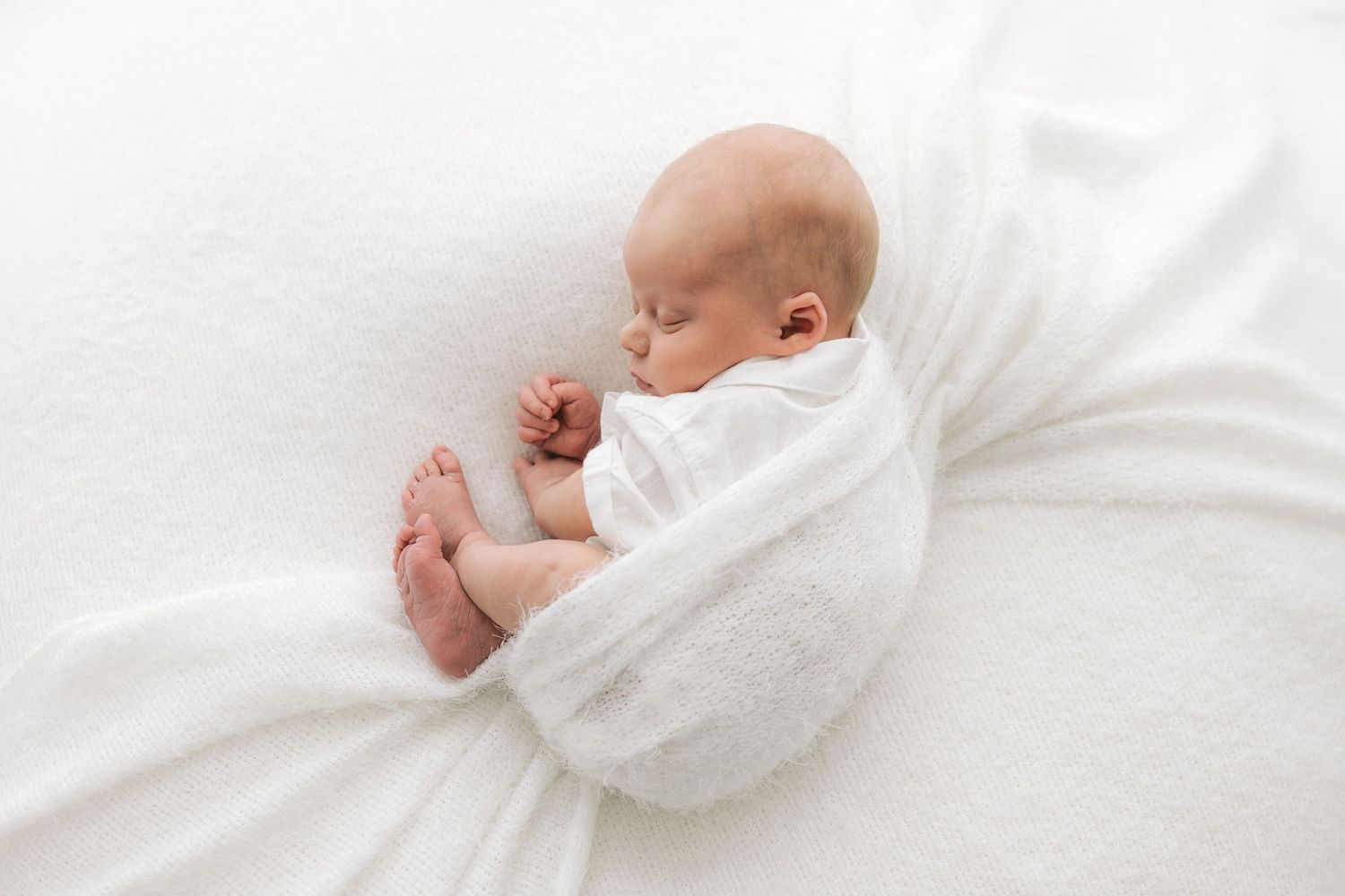 tiny newborn baby is curled up in a white outfit against a white background, sleeping soundly in natural newborn photoshoot in Ayrshire