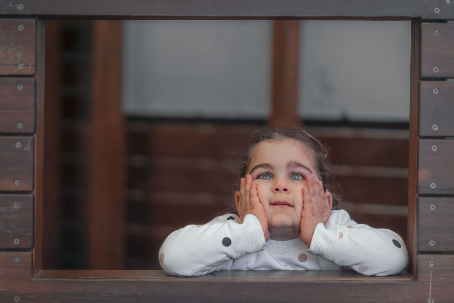 A child in a white sweater gazes upward through a window frame with hands resting on their cheeks.