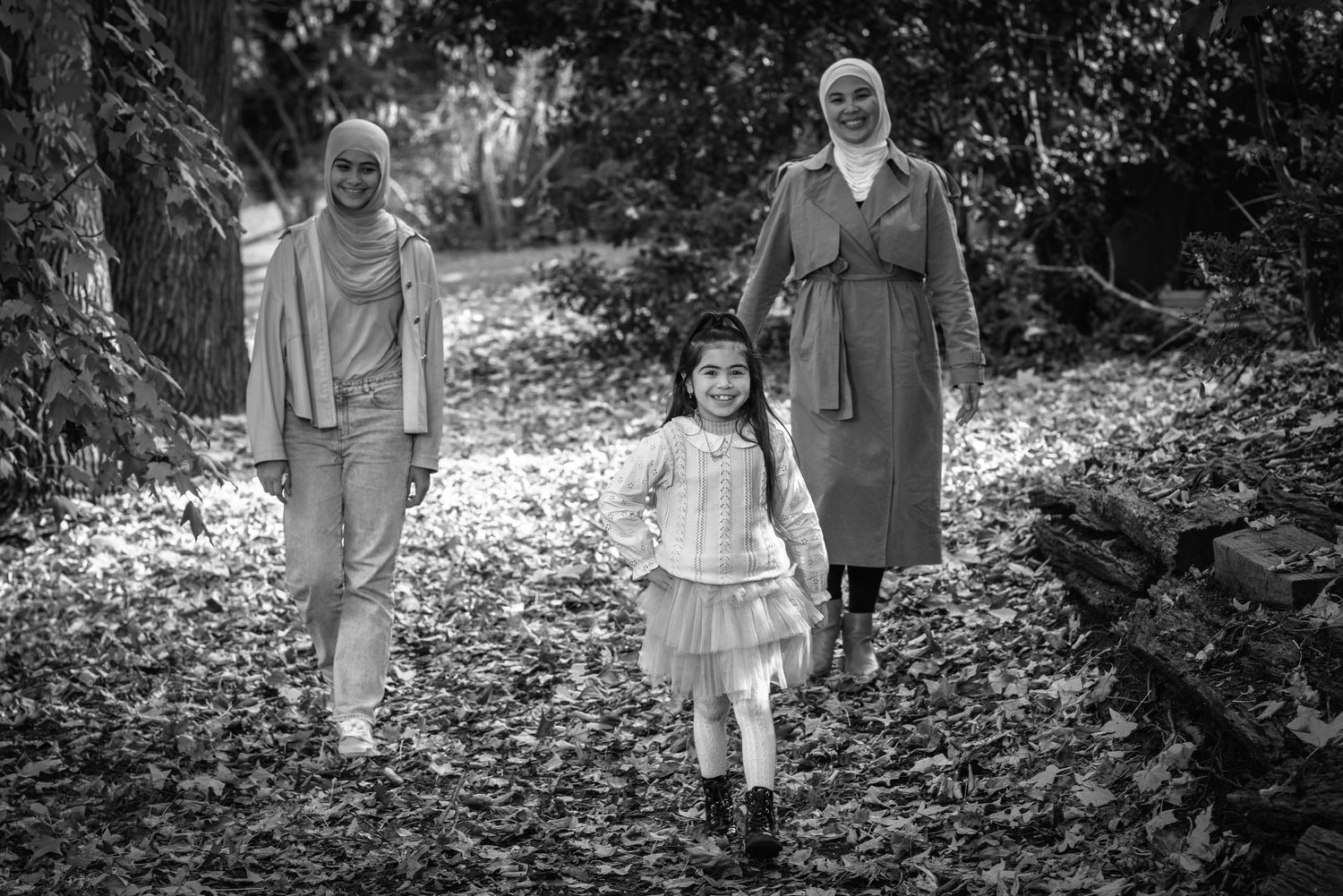 Three people walking together through fallen autumn leaves on a forest path in black and white.