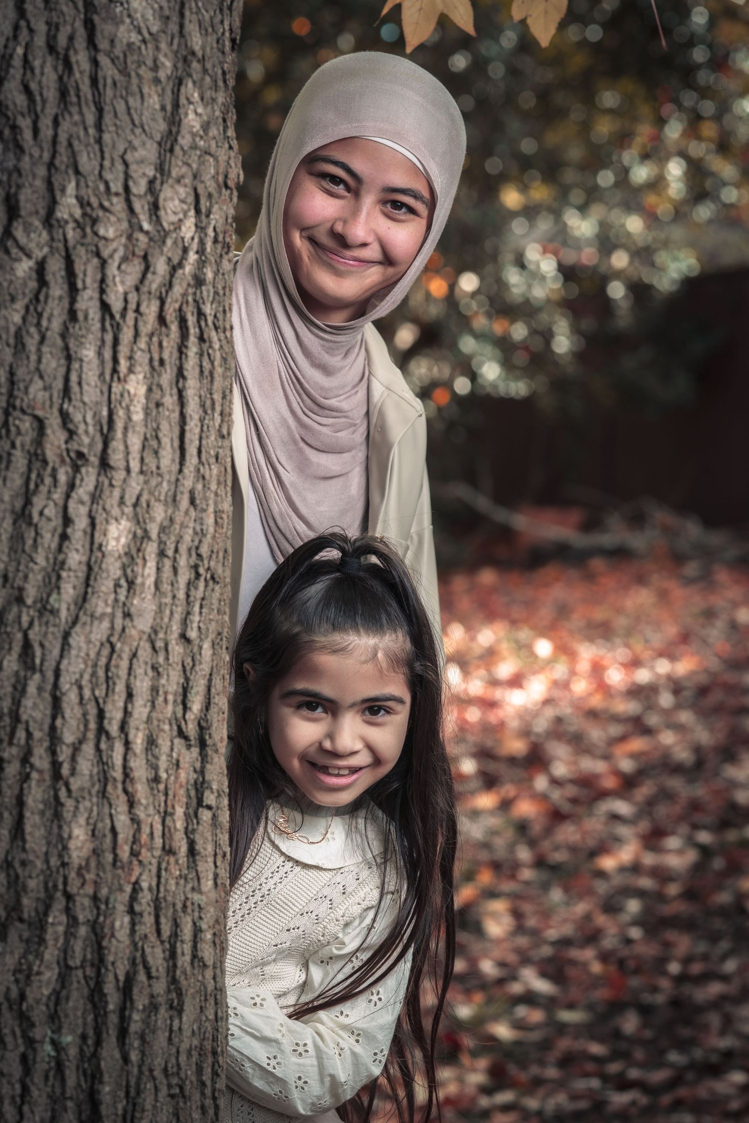 Two people playfully peeking from behind a large tree trunk in an autumn forest setting.