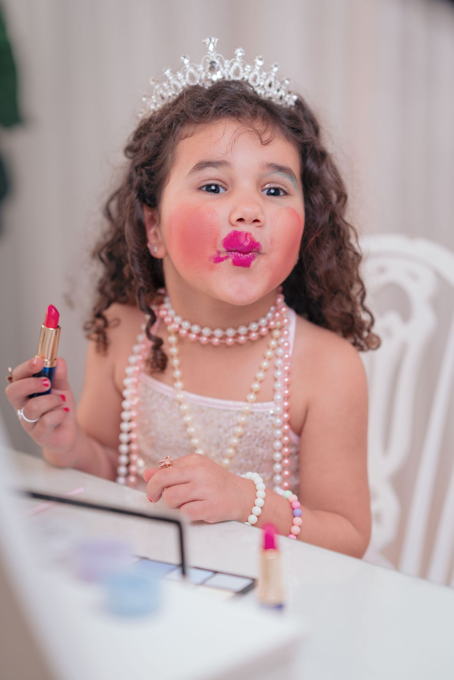 A series of photos showing a child wearing pearl necklaces playing with pink lipstick at a white table.