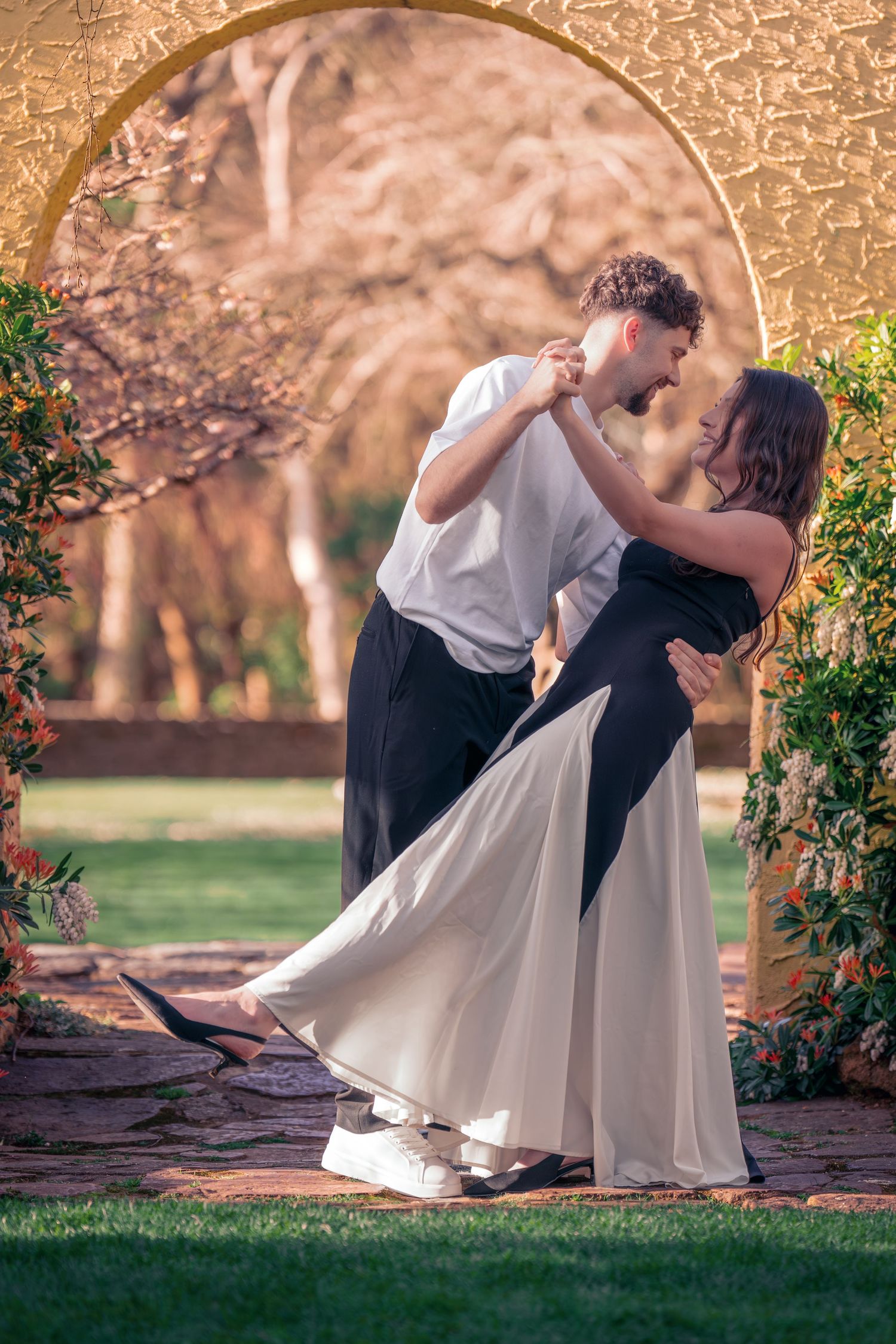 A romantic couple dancing in a garden archway with blooming flowers and soft evening light.