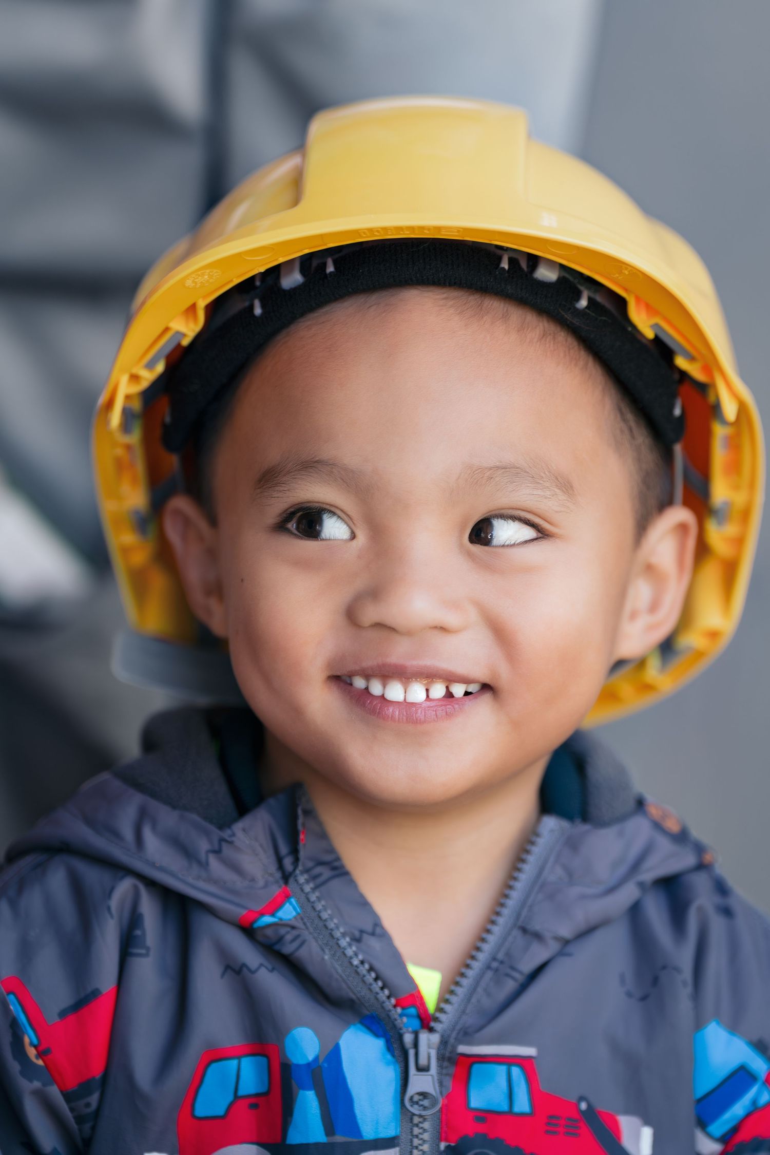 Child wearing yellow hard hat and blue jacket smiling with a playful expression.