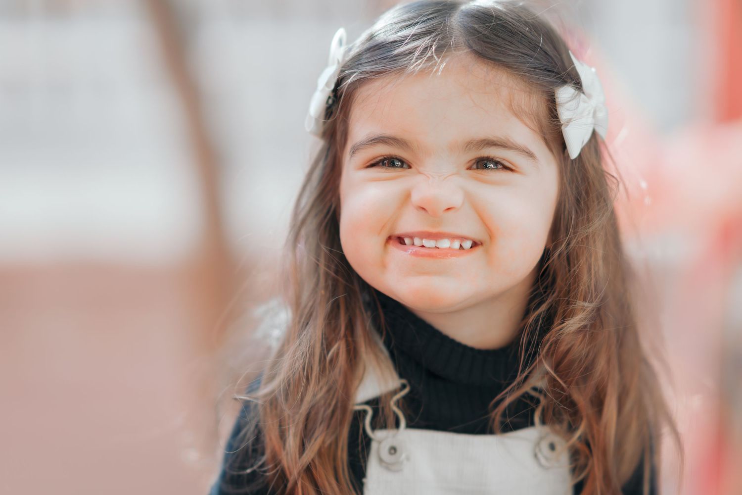 A joyful child wearing a black turtleneck and white overalls with bows in their long dark hair.