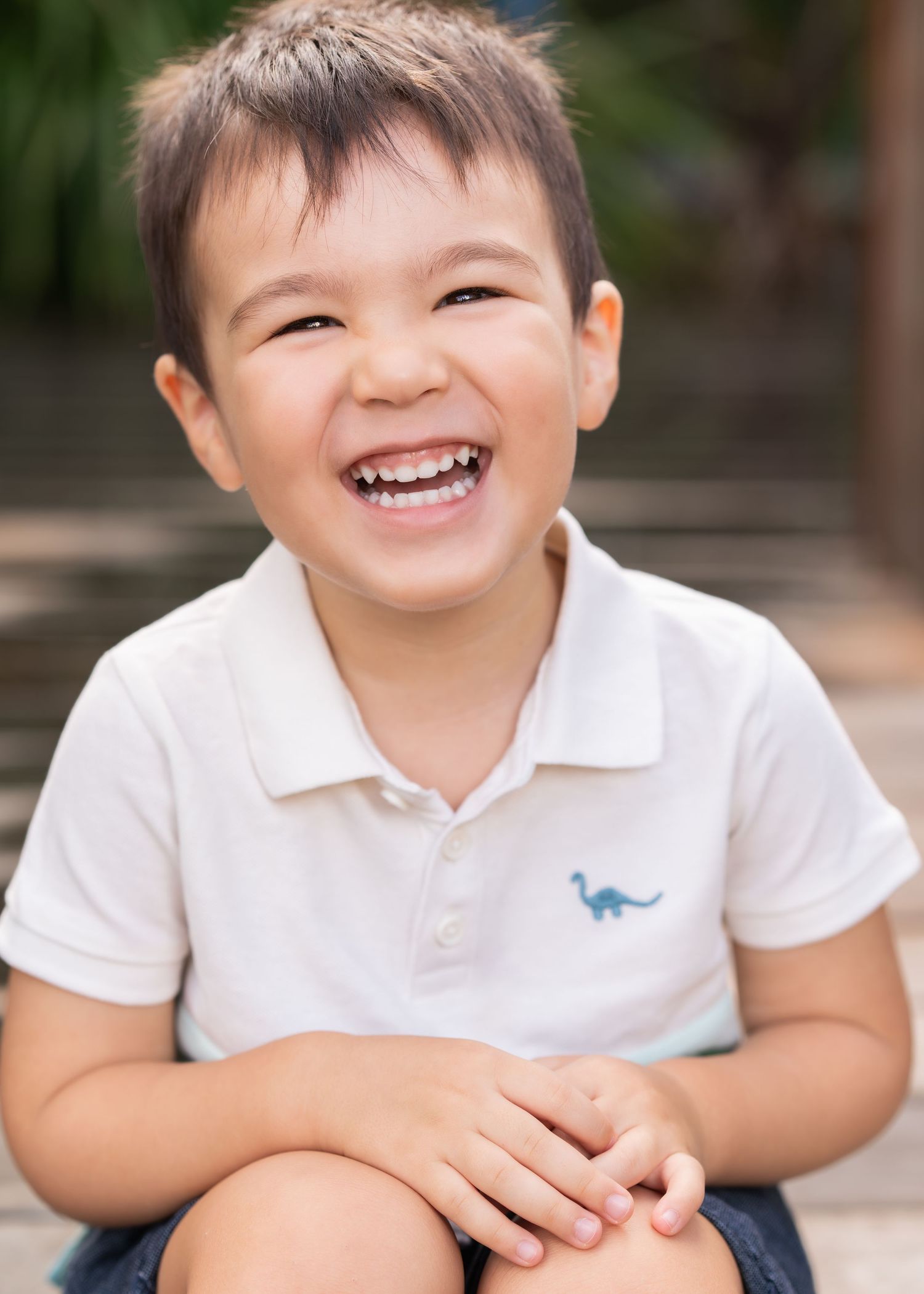 Adorable young boy in a white polo shirt with a dinosaur logo laughing joyfully outdoors.