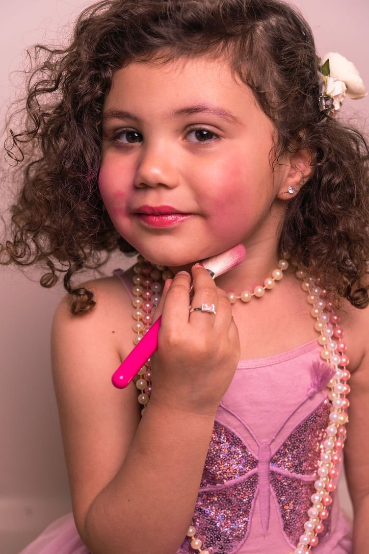 A curly-haired child wearing pink and pearls poses elegantly with sparkly dress details.