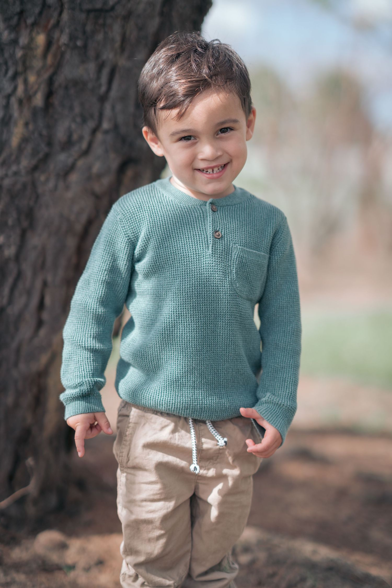 Young child in mint green sweater smiling while standing next to tree trunk outdoors.