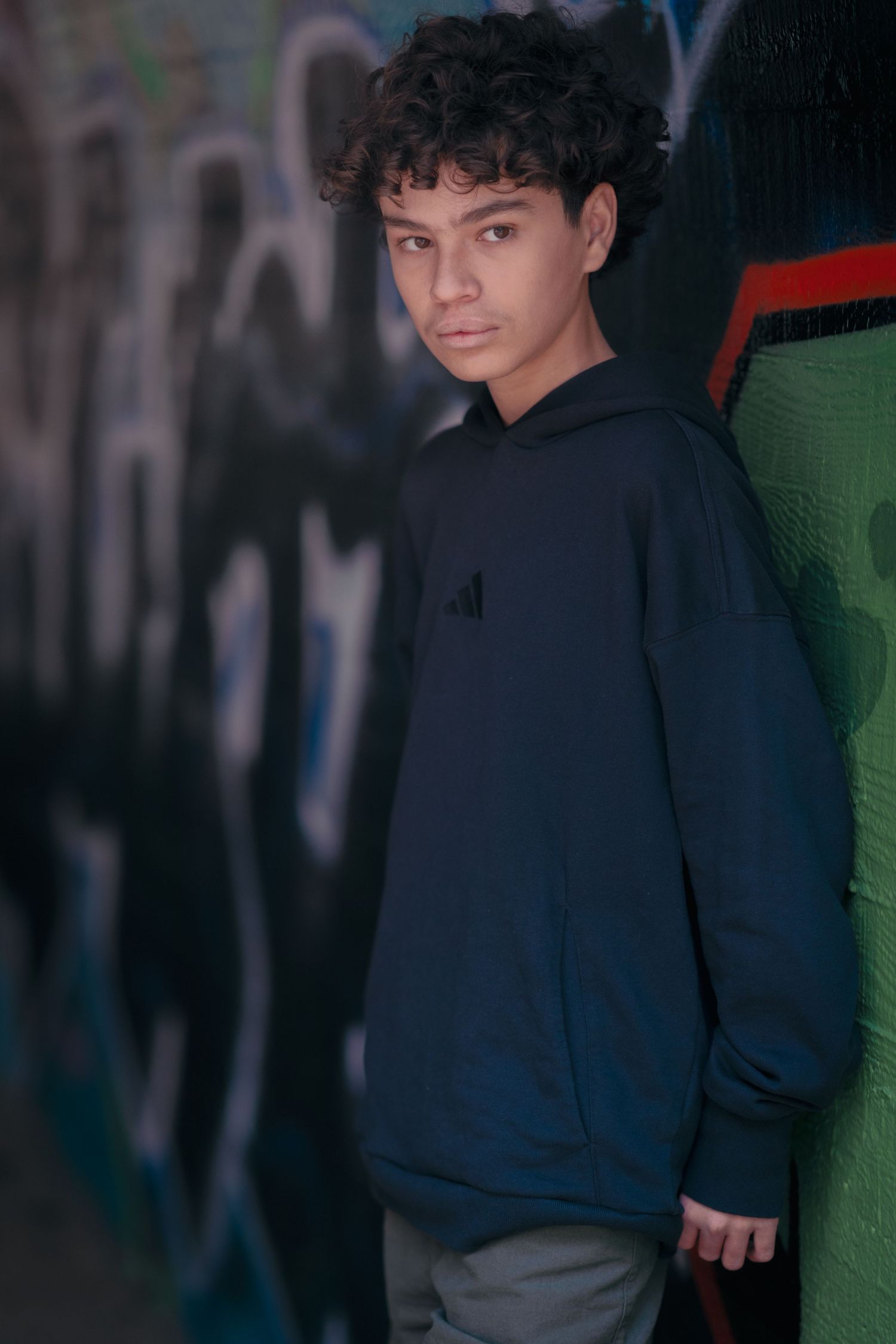 Person in black hoodie posing against colorful graffiti wall in moody lighting.