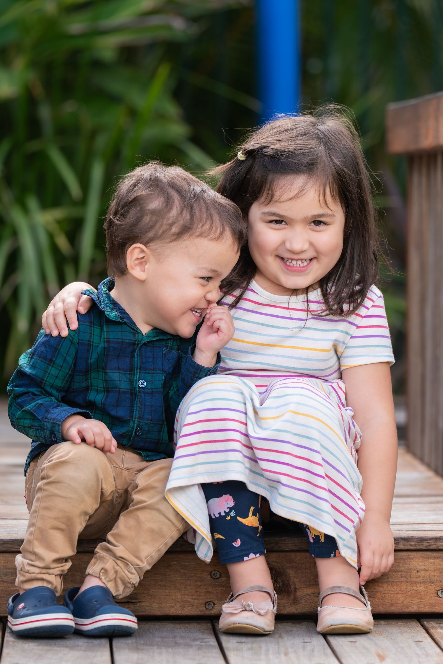 Two young siblings sitting together on wooden steps, smiling and hugging outdoors.