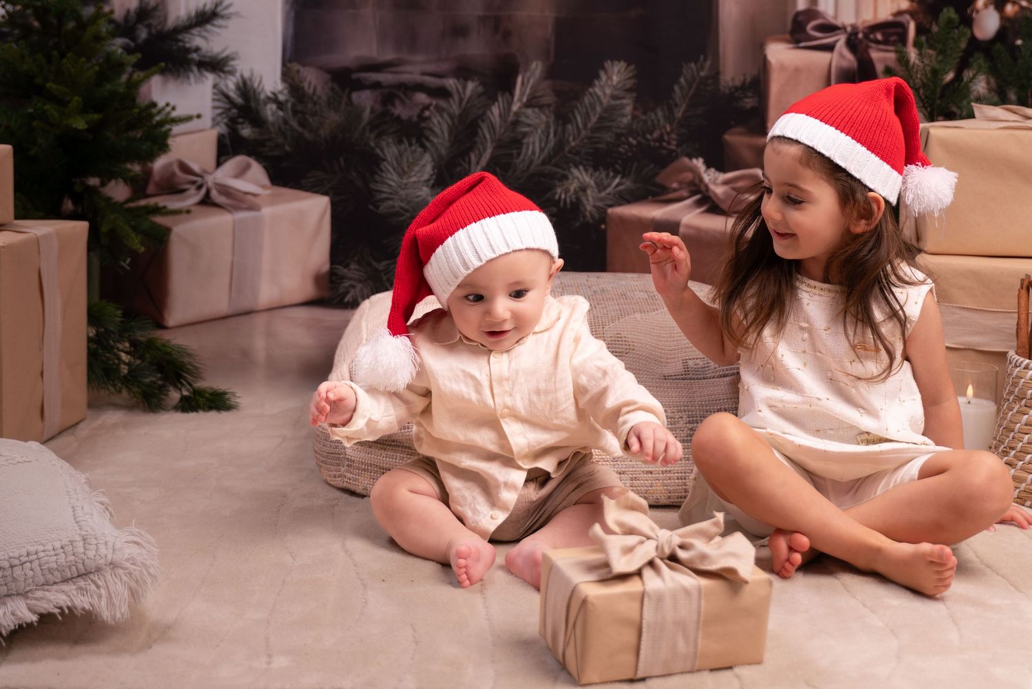 Two children in Santa hats sit on the floor near wrapped presents and a Christmas tree in a cozy holiday setting.