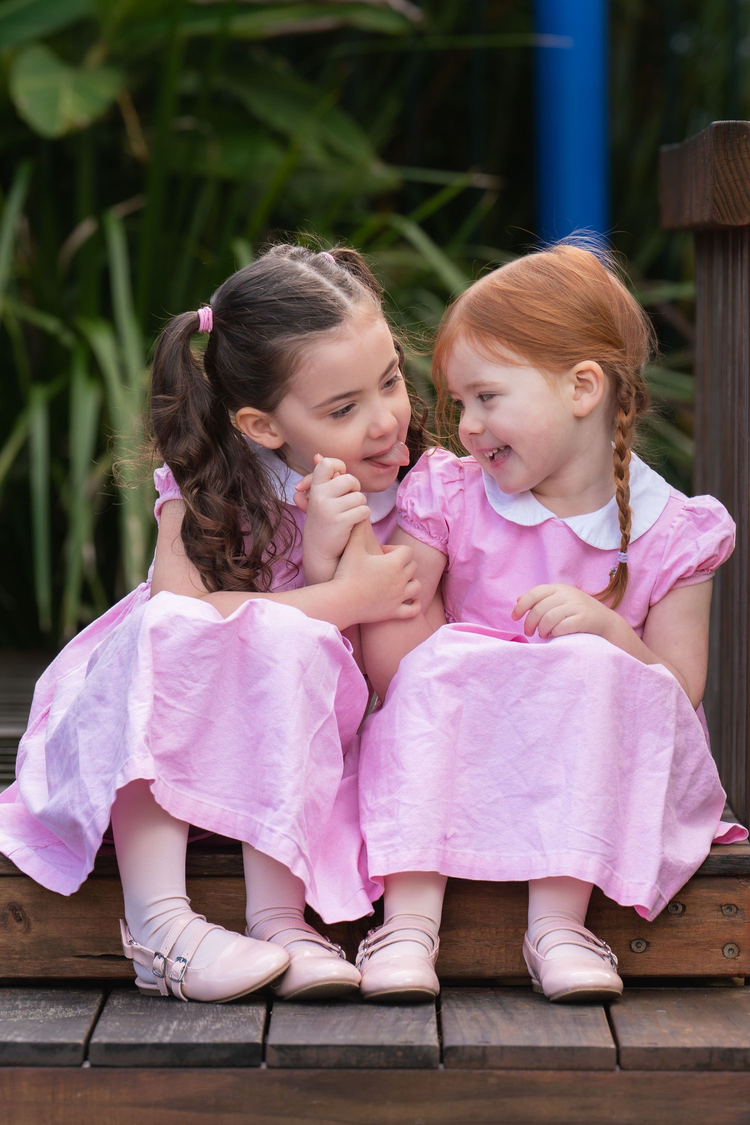 Two young girls in matching pink dresses sitting together on wooden steps, smiling and laughing outdoors.