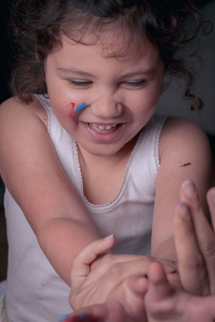 A child with curly hair and colorful face paint smiles playfully while reaching forward.