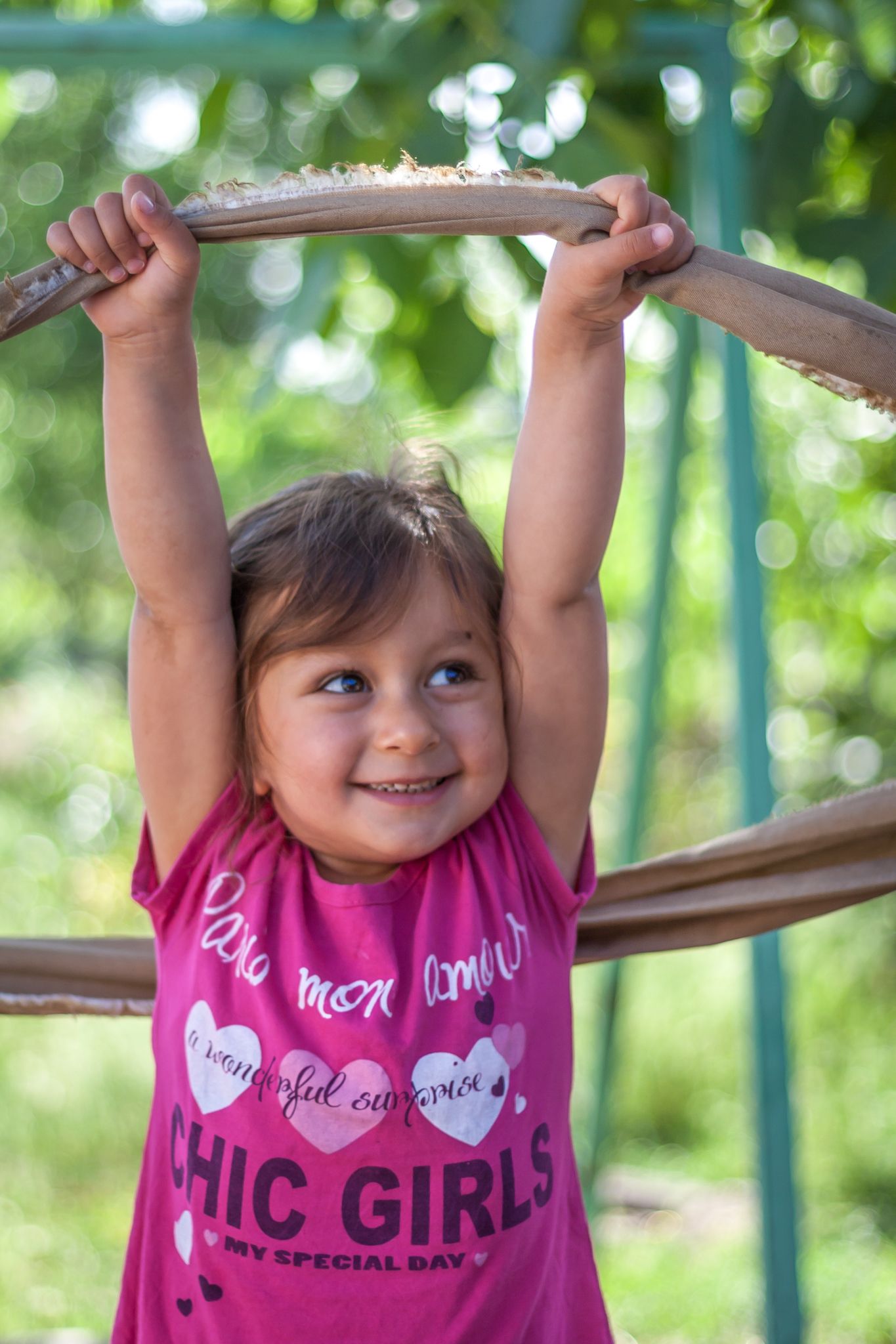 A child in a pink shirt reaches up to grab playground equipment with a joyful expression.