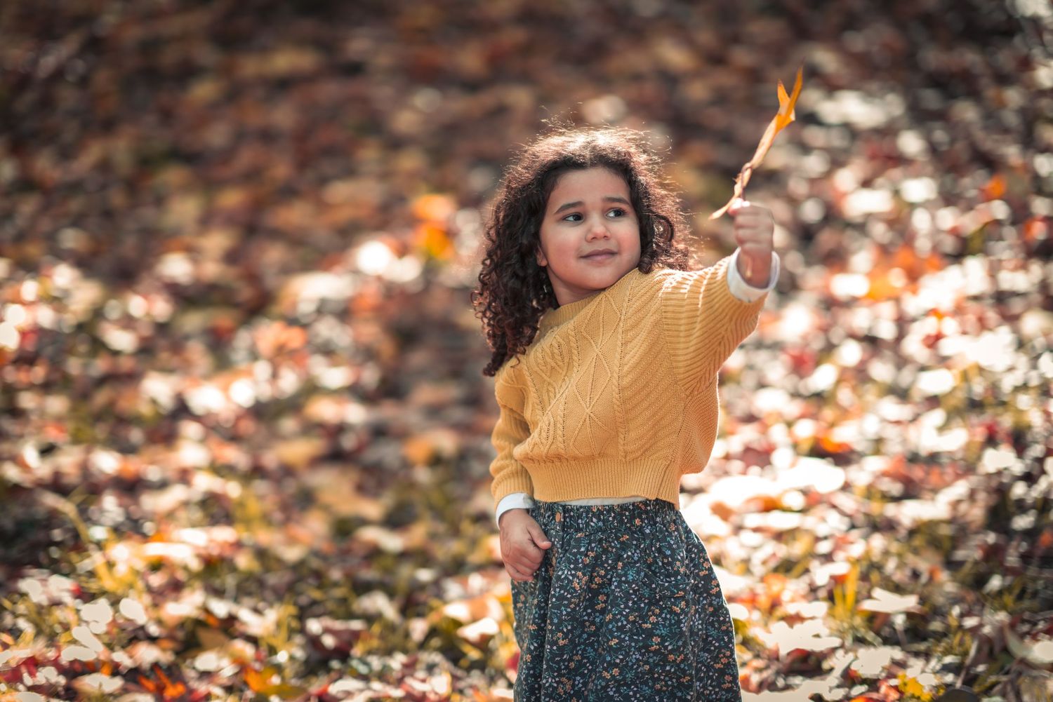 A child in a mustard sweater points upward while standing among fallen autumn leaves.