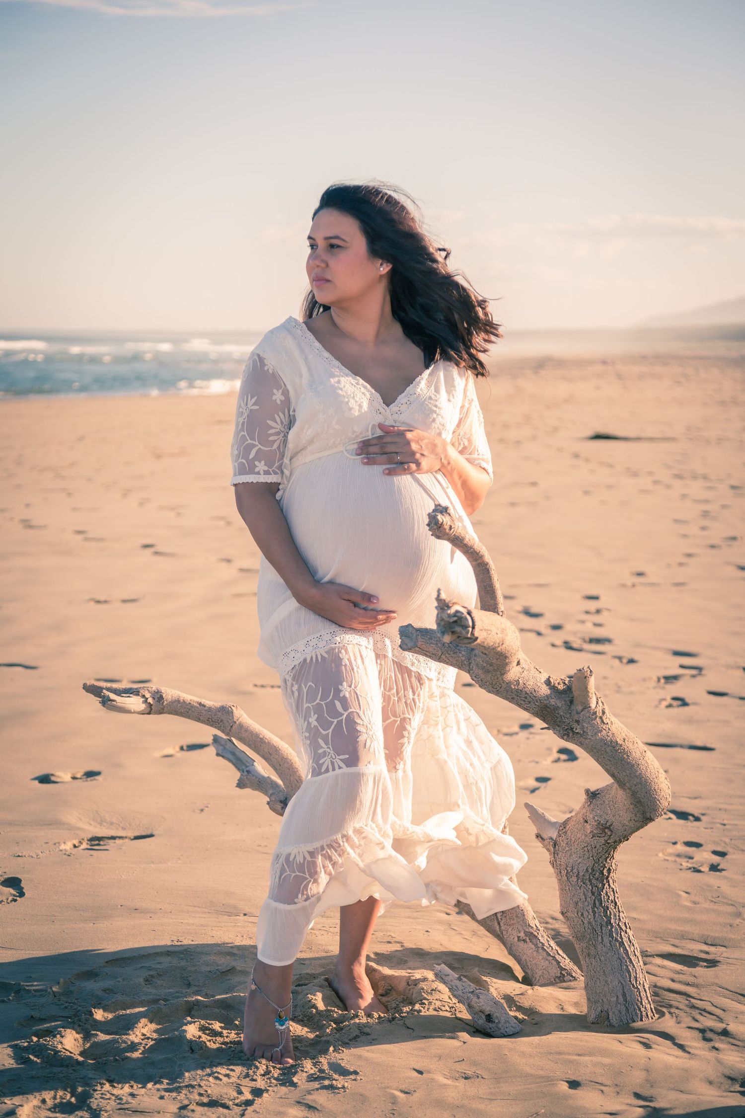 A flowing white lace dress billows in the breeze next to driftwood on a peaceful beach.