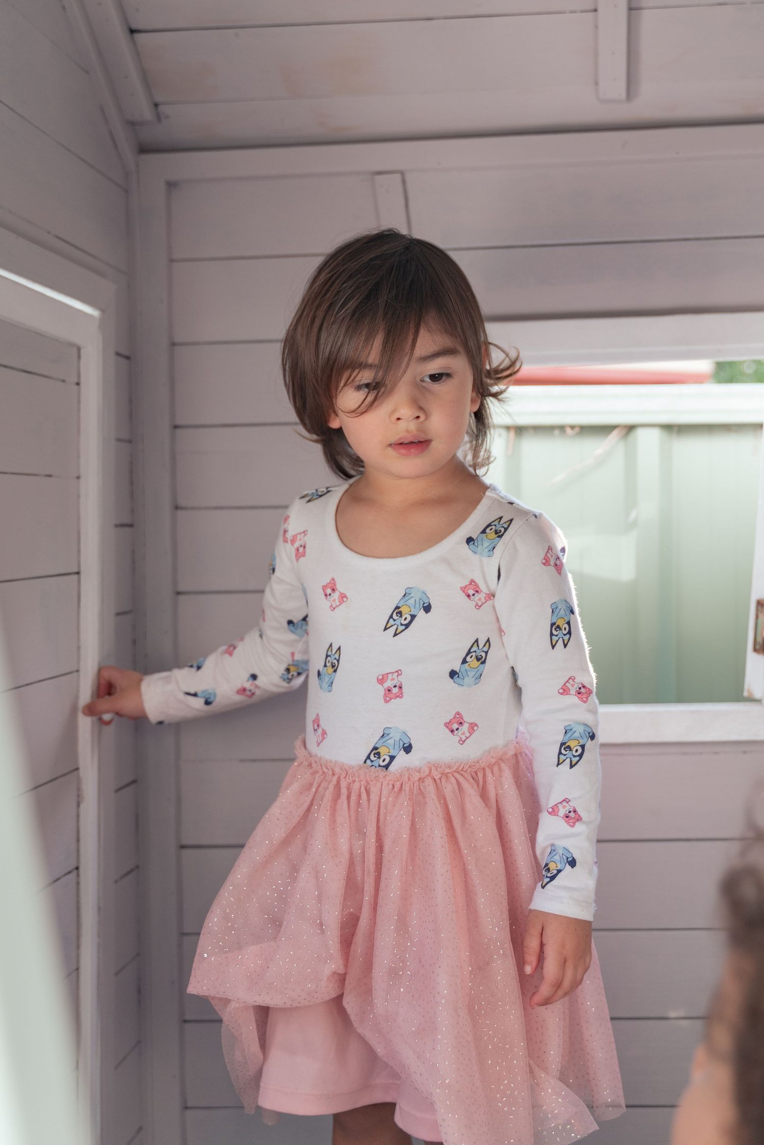 A child wearing a dress with toy print stands in a white wooden playhouse.