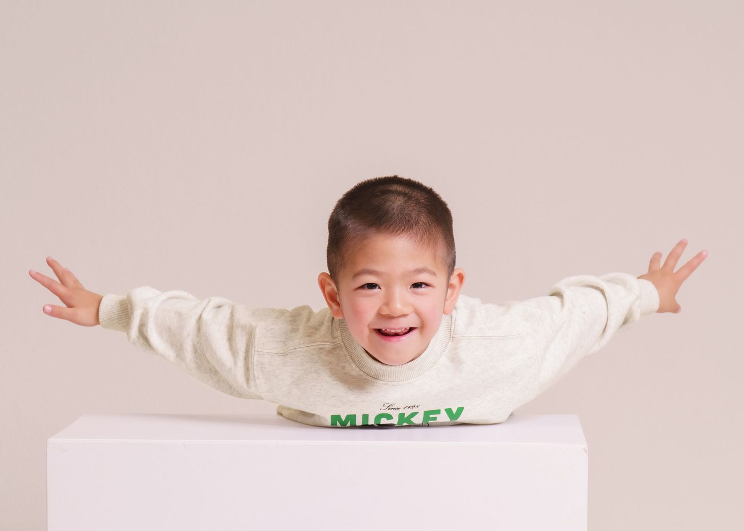 Happy toddler boy in Mickey Mouse sweater spreading arms wide, smiling joyfully against neutral background.