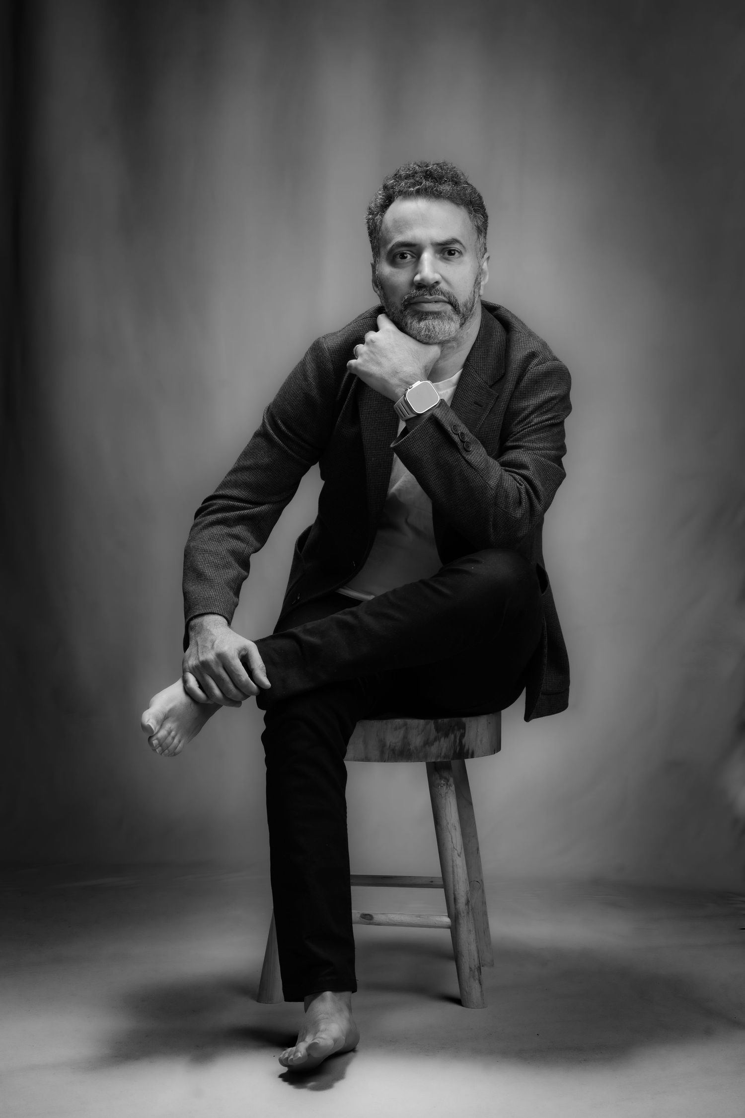 Artistic black and white portrait of someone seated on a wooden stool in a dark suit against a shadowy background.
