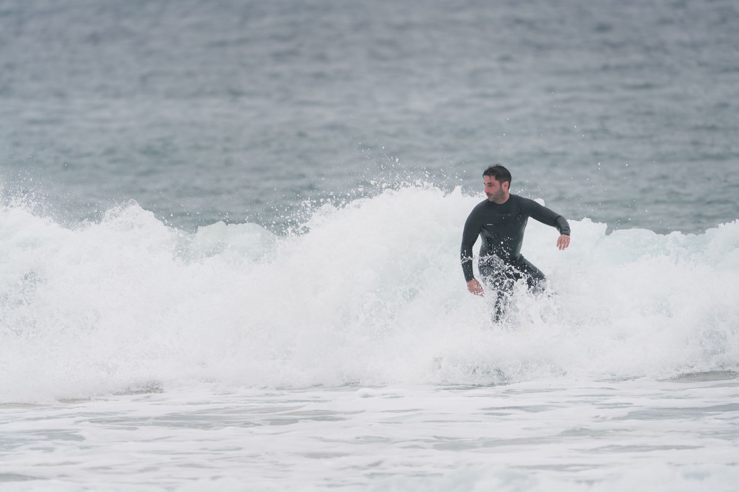 Surfer in black wetsuit catching a wave on a grey overcast day.