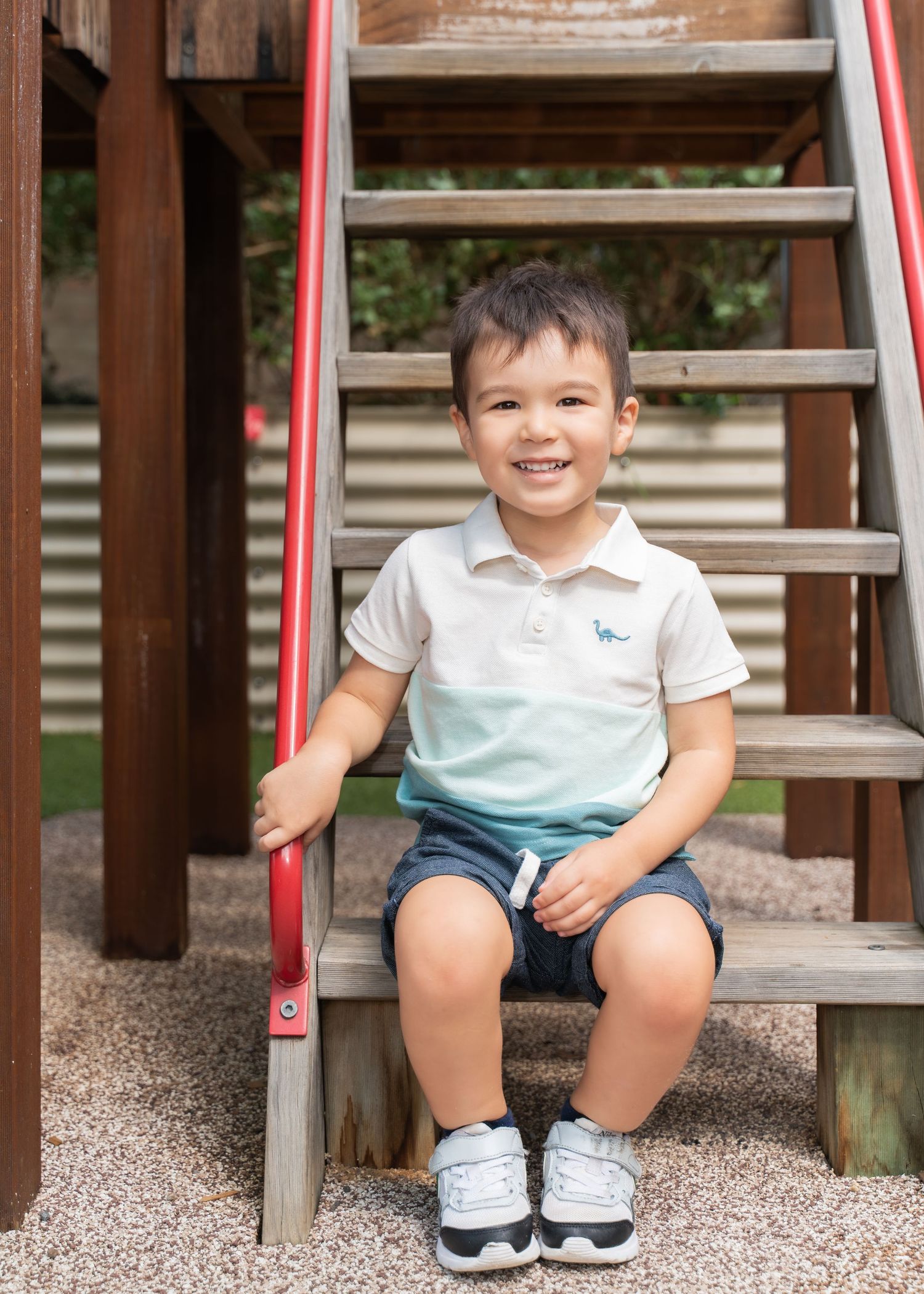 Smiling toddler boy sitting on playground steps, wearing a white polo shirt and navy shorts, holding a red railing.