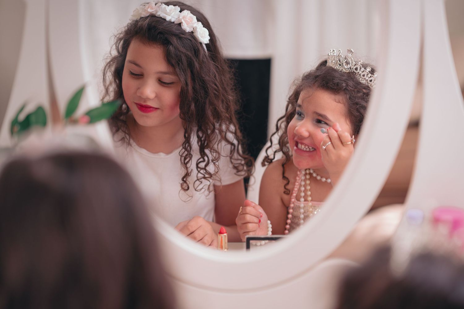 Two people wearing tiaras smile while looking into an oval mirror during a special occasion.