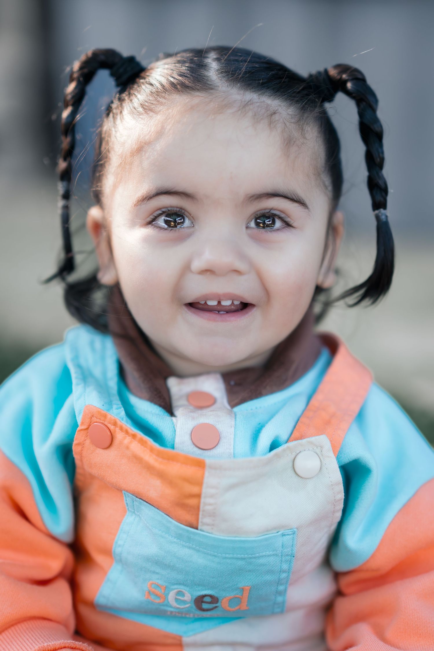 Baby with pigtails wearing turquoise and orange outfit smiling brightly at the camera.