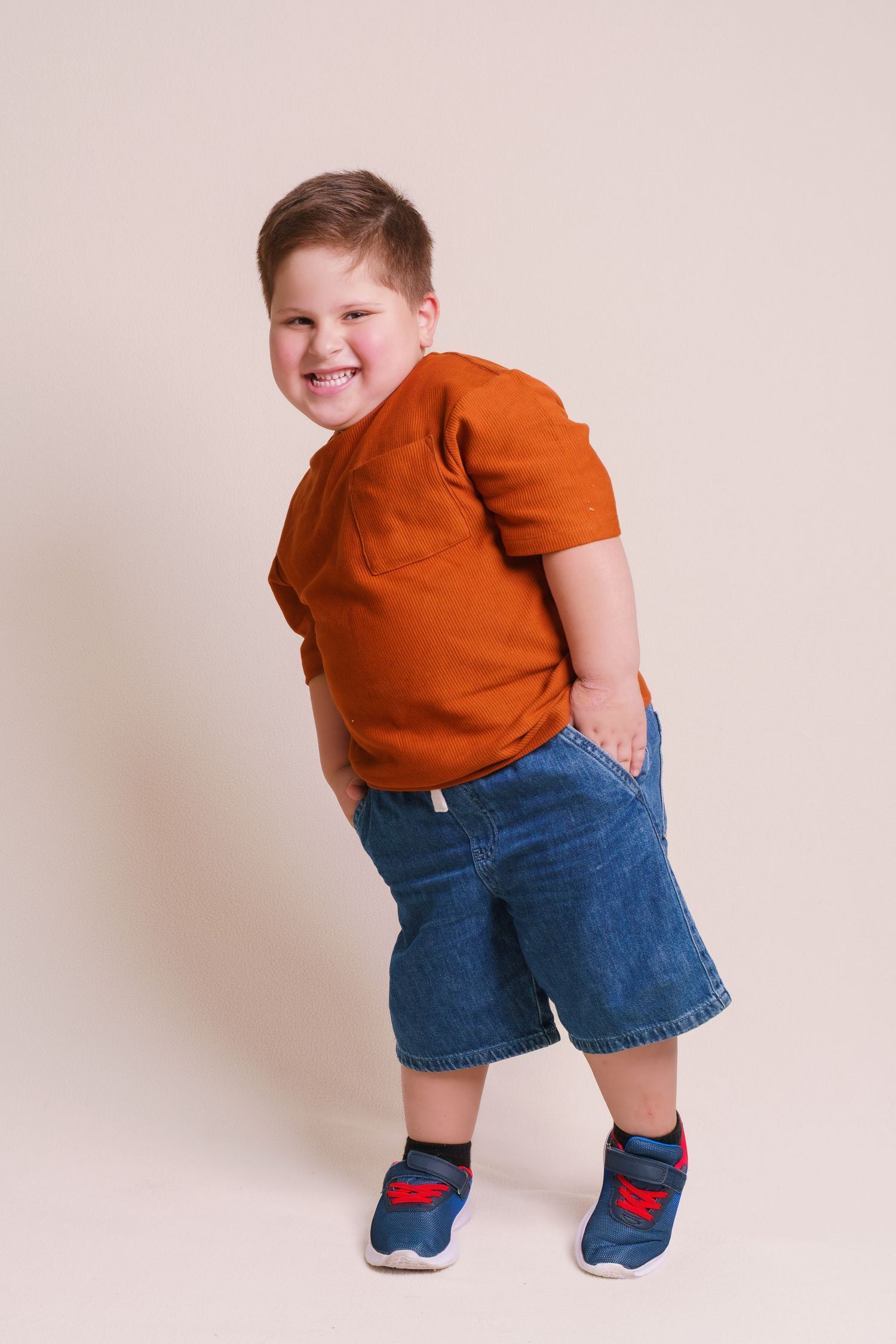 Smiling chubby boy in an orange t-shirt and denim shorts posing playfully against a beige background.
