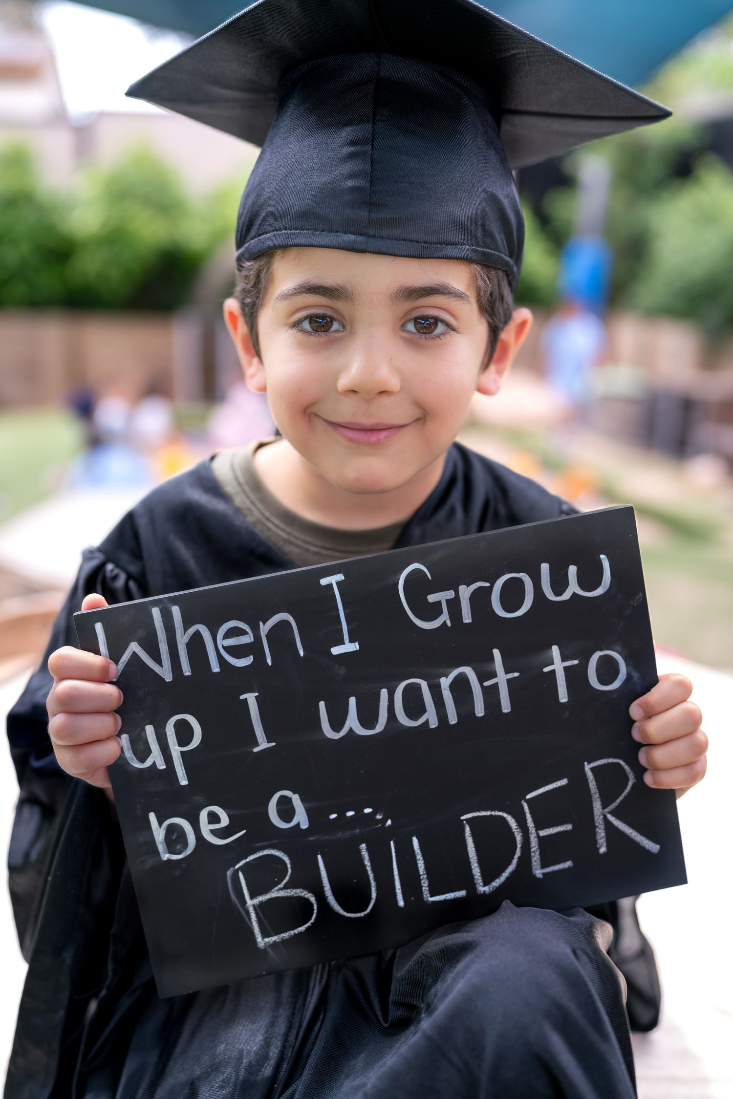 A child in graduation cap holds a chalkboard sign with the message 'When I grow up I want to be a builder'.