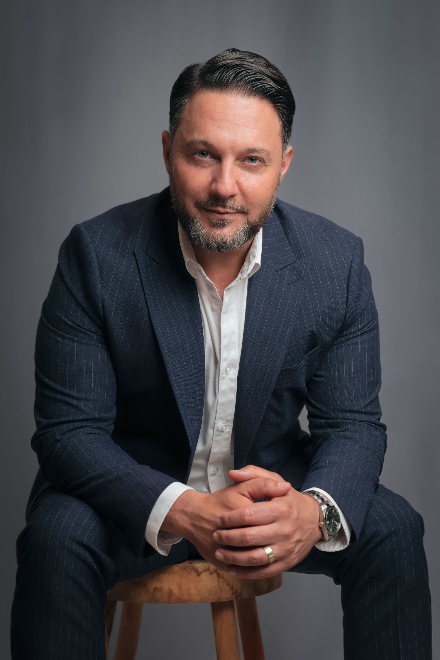 Man in navy pinstripe suit seated on wooden stool, hands clasped, professional headshot on gray background.