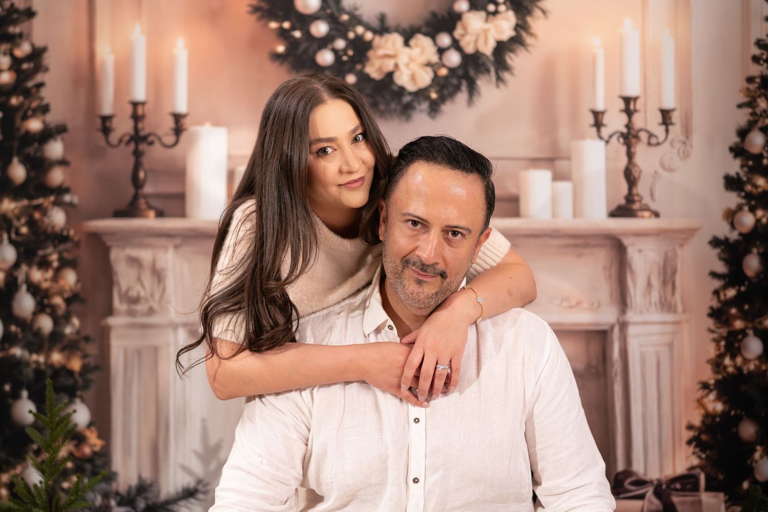 A romantic couple poses in an elegant holiday setting with white decorations and candles in the background.