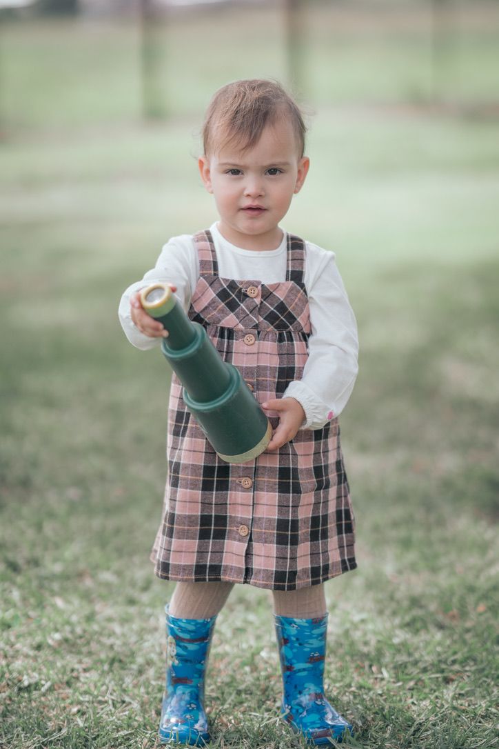Child in plaid dress and rain boots holds green thermos while standing in grassy field.