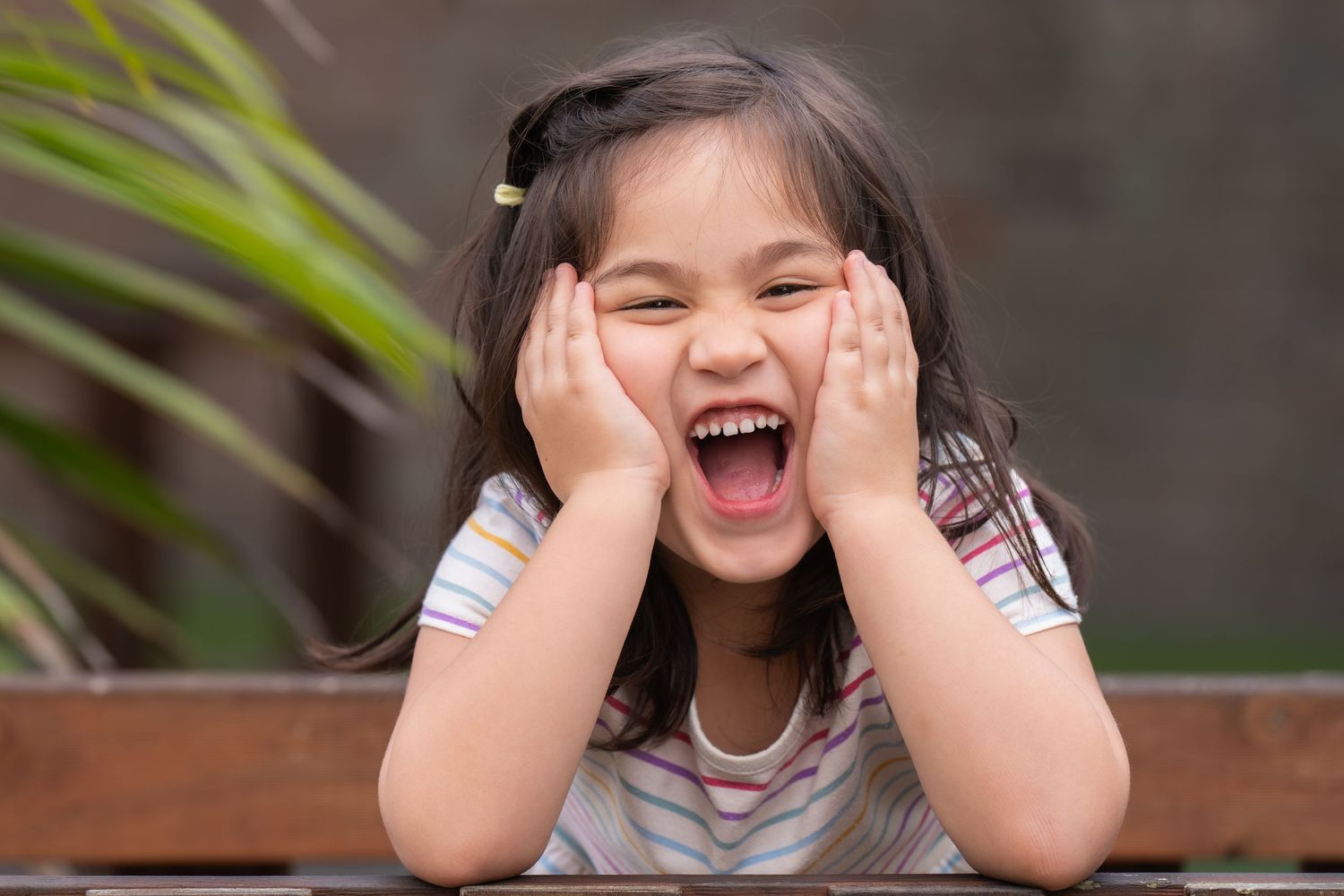 Excited young Asian girl laughing with hands on cheeks, sitting at a wooden table outdoors.