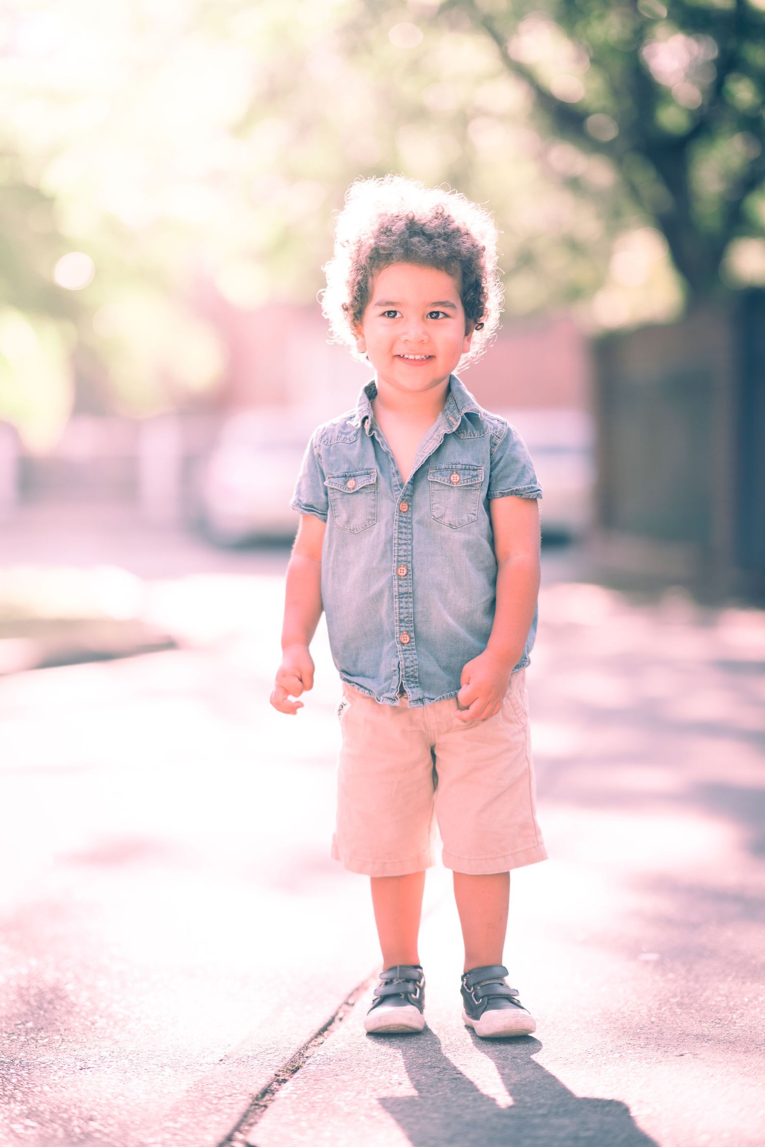 A child wearing denim and pink shorts stands on a sunny sidewalk.
