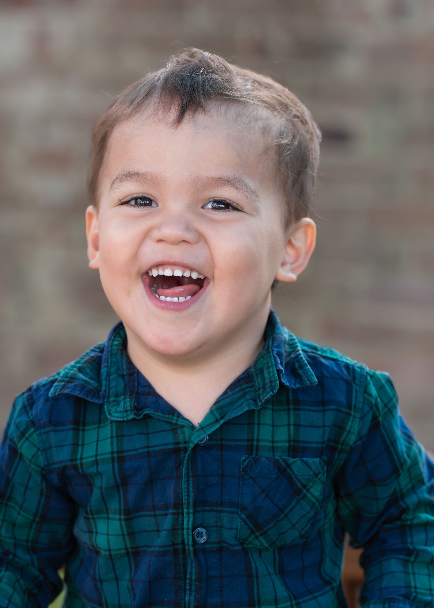 Smiling toddler boy in green plaid shirt laughing outdoors with brick wall background.