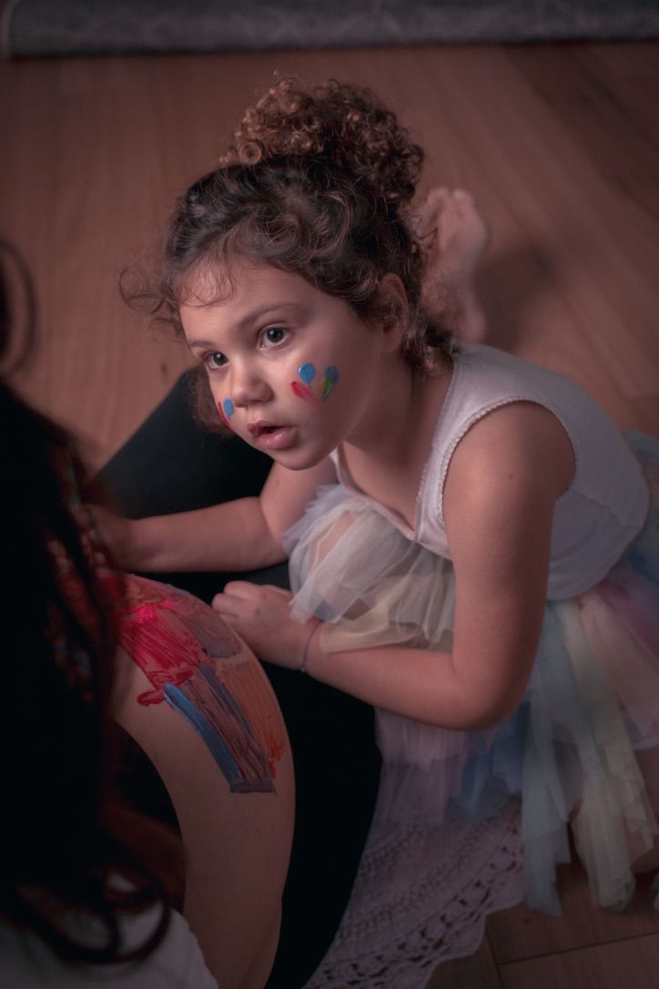 A child in a white dress with face paint leans over to draw colorful artwork on someone's leg.