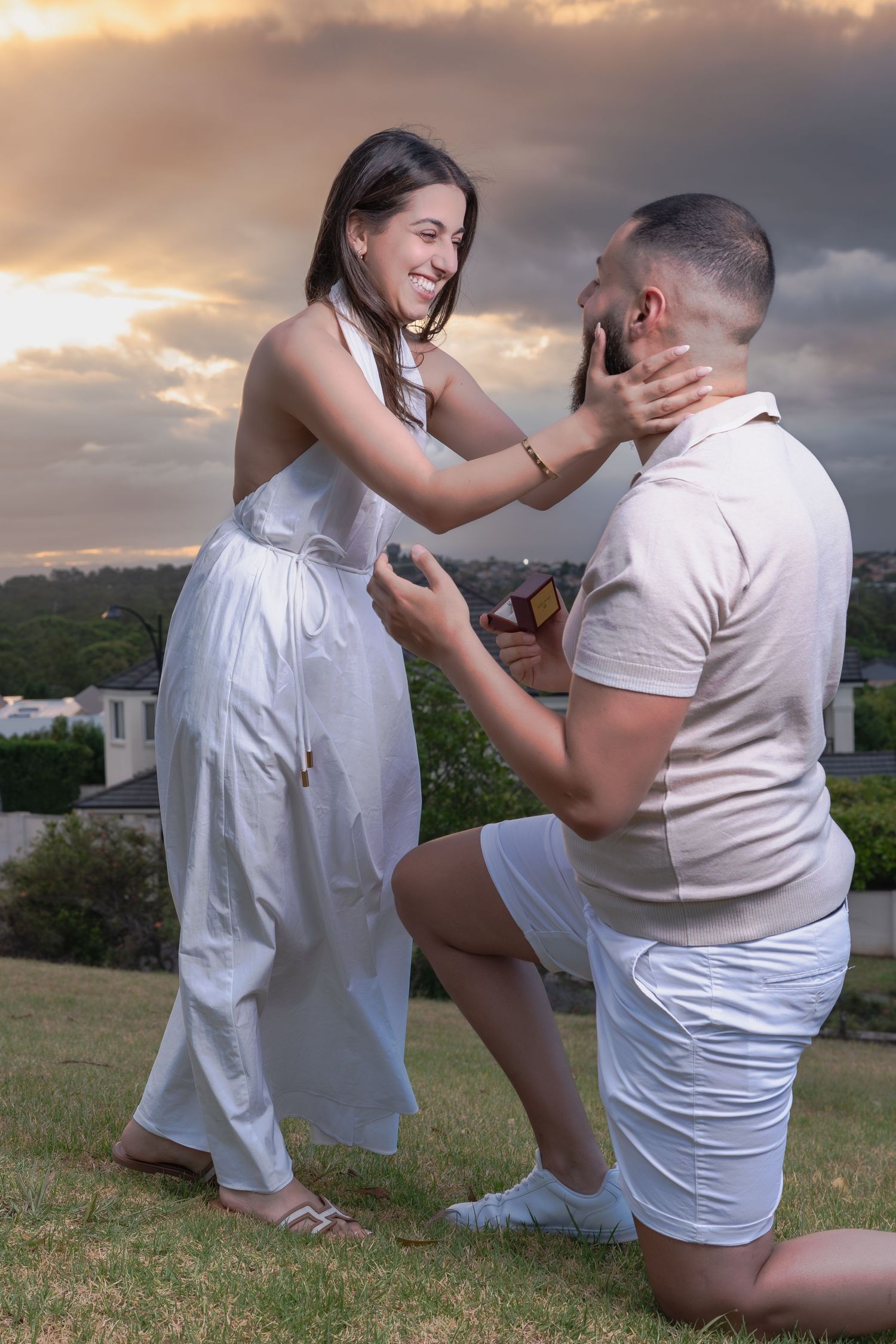 A romantic proposal scene unfolds at sunset with someone in a white dress and another in a white shirt kneeling on grass.