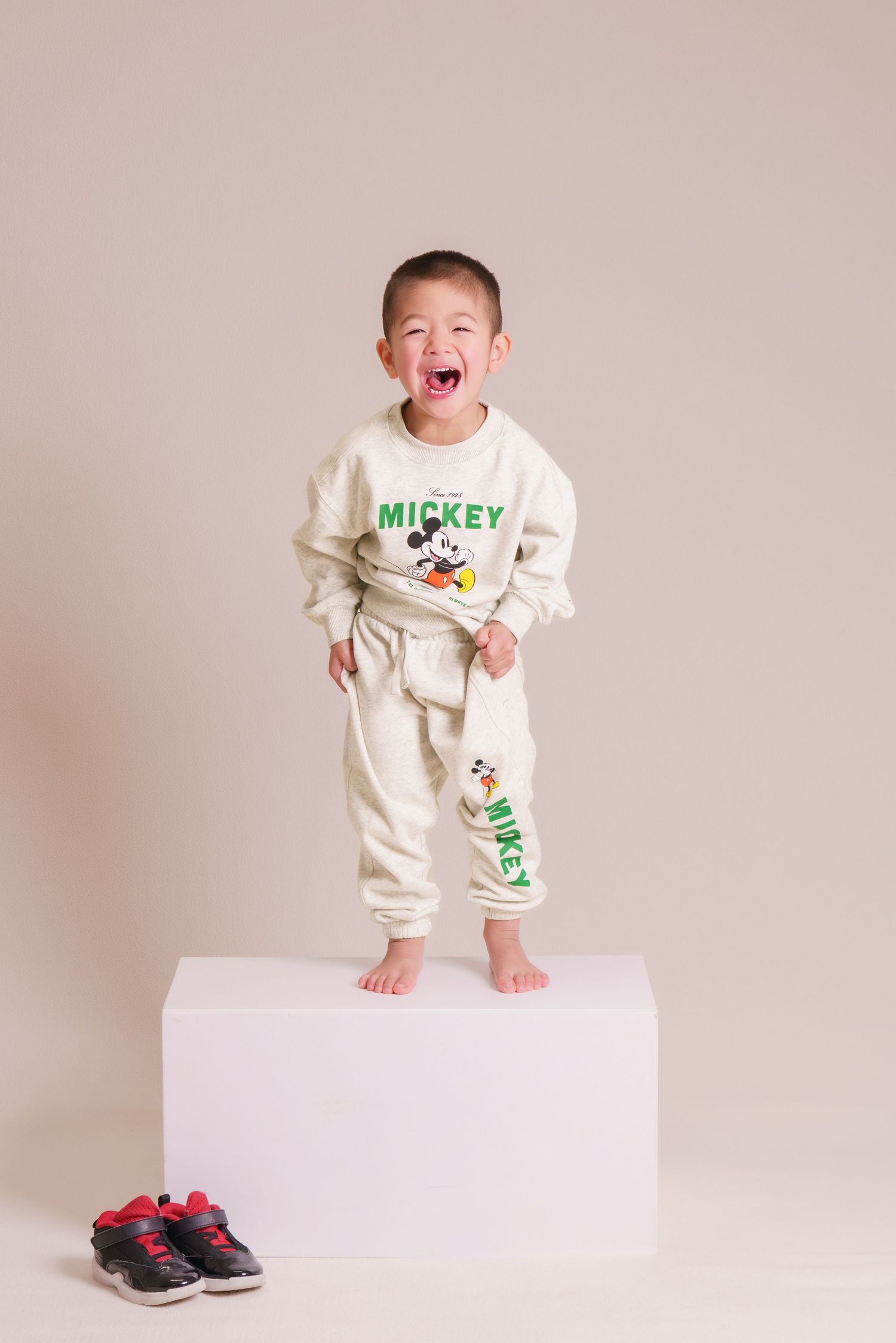 Joyful toddler boy wearing Mickey Mouse tracksuit standing barefoot on white block, shoes beside him.