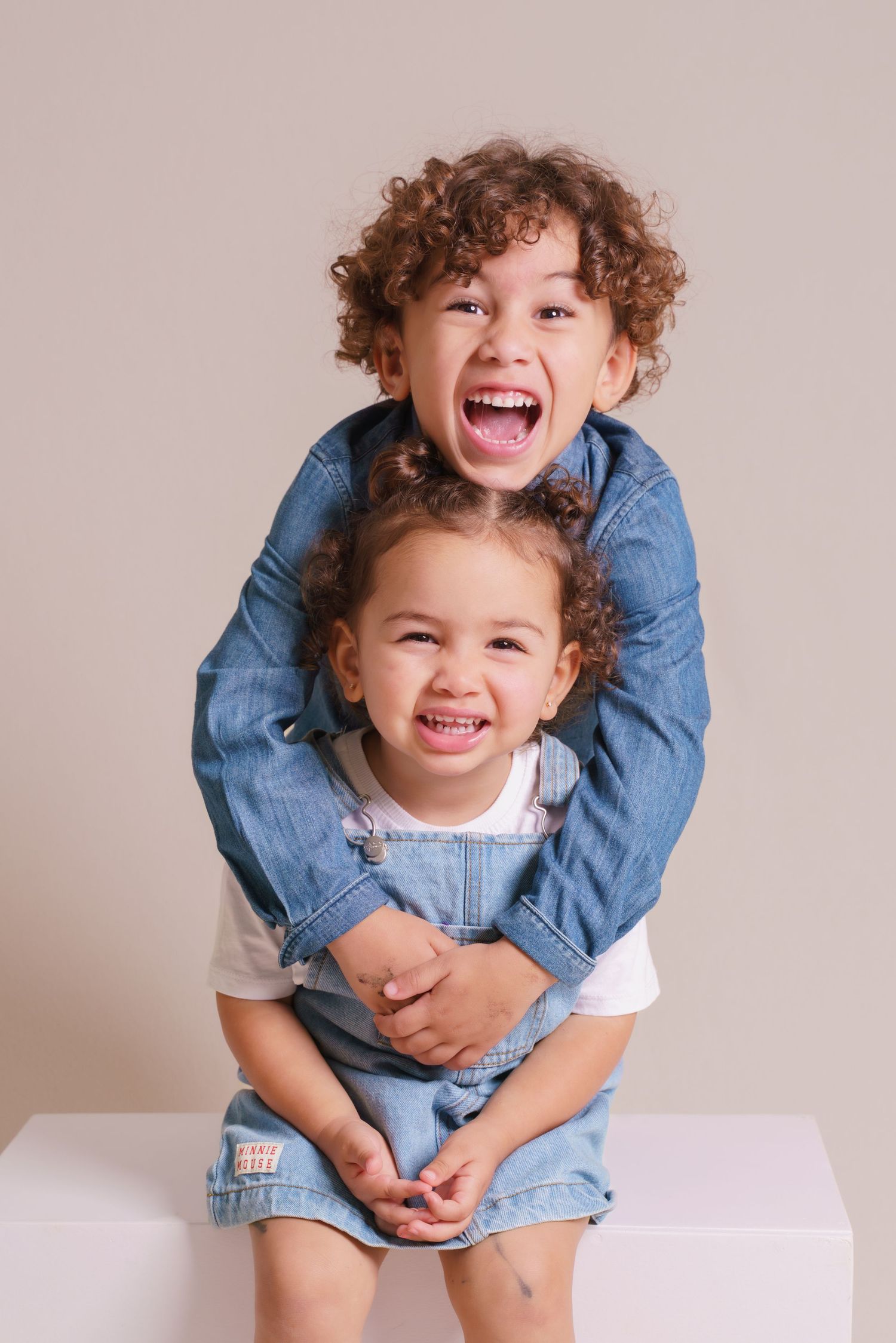 Two joyful young siblings in denim outfits hugging and laughing together against a neutral background.