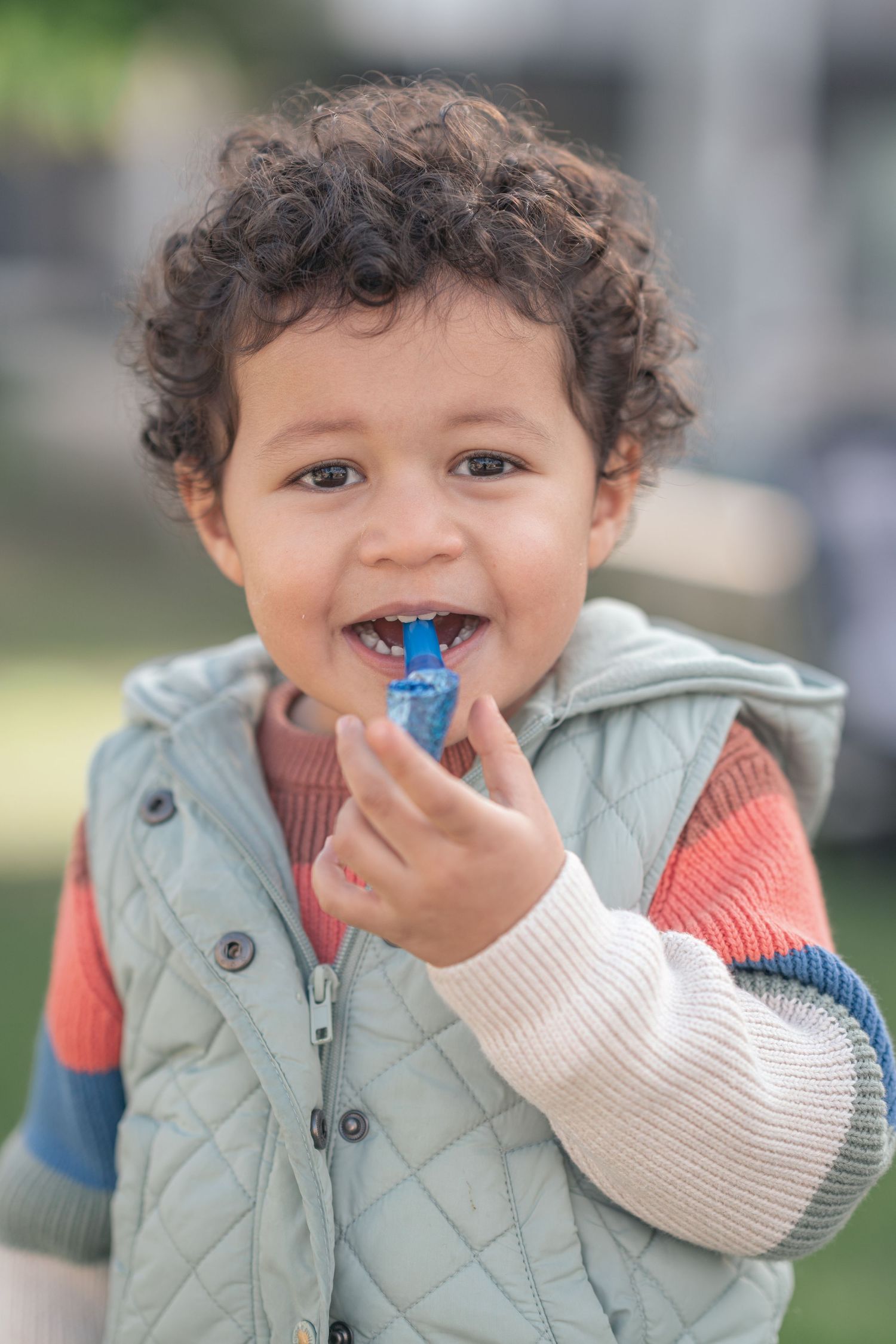 A young child in a quilted vest smiles while holding a blue toy outdoors.