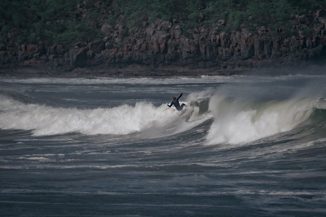 Surfer performs aerial maneuver on large ocean wave against rocky cliff backdrop.
