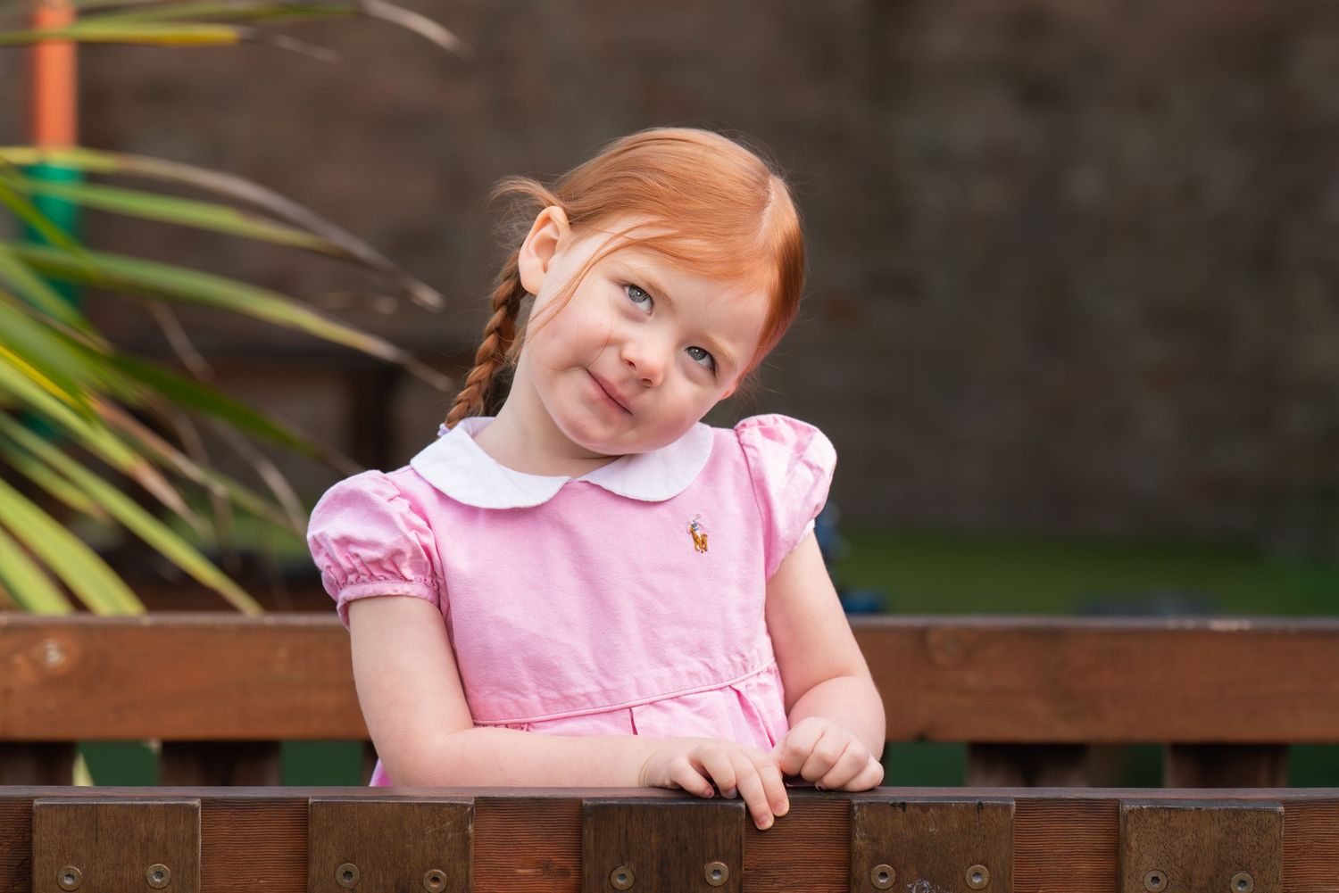 A cheerful red-haired girl in a pink dress leans on a wooden fence, smiling outdoors.