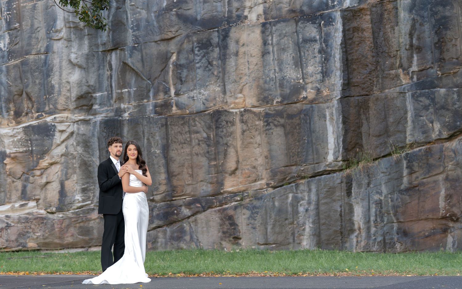 Wedding couple poses against dramatic rocky cliff face in outdoor setting.