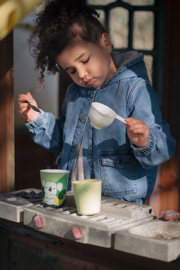 Young child in denim jacket carefully pouring ingredients while playing at an outdoor counter space.