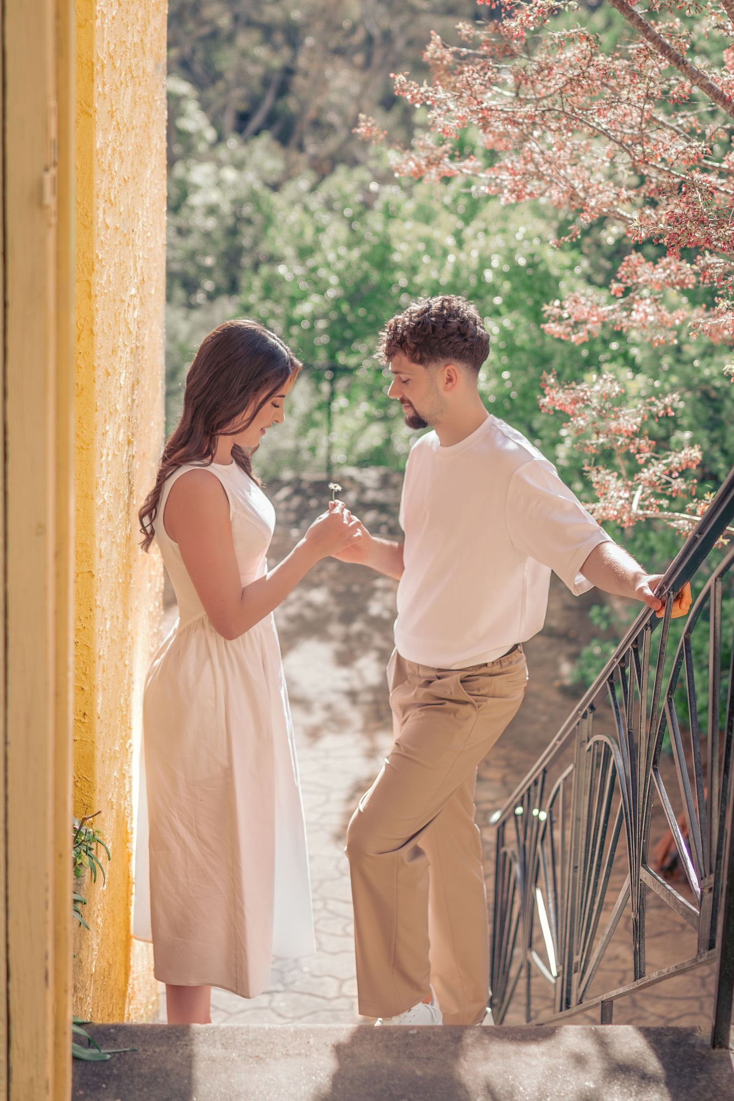 Couple in light colored outfits standing together on a balcony with flowering trees in the background.